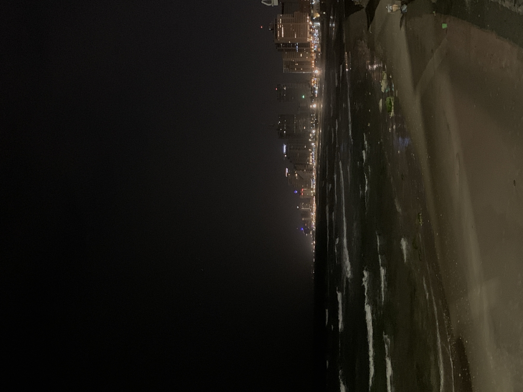 The image shows a night view of the Tel Aviv, Israel coastline. Tall buildings with illuminated windows are visible in the distance, creating a city skyline. The dark ocean is in the foreground, with visible waves reflecting the city lights. A walkway or sidewalk runs along the beach, appearing damp, with indistinct dark figures present on it.
