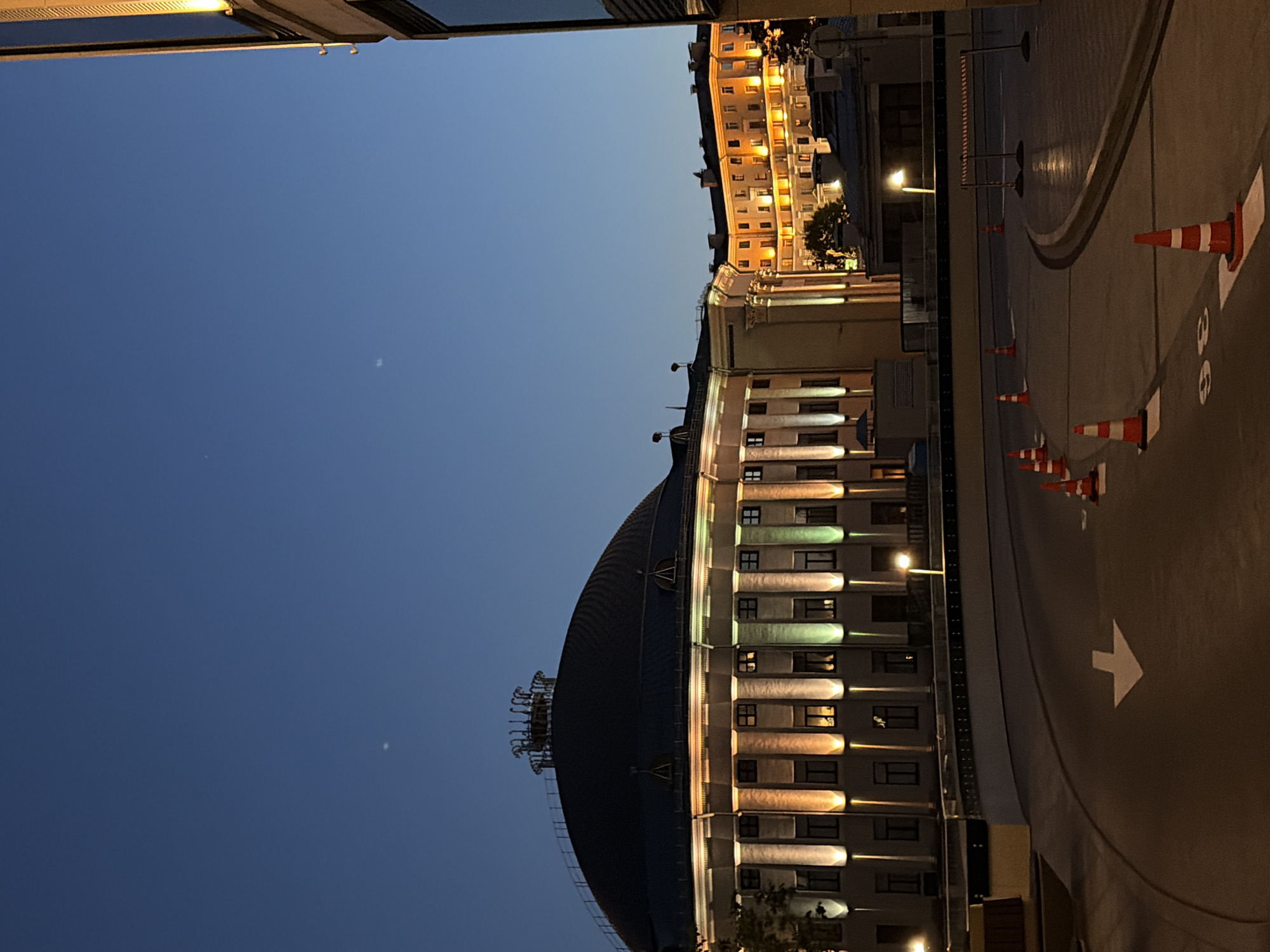 This is a vertical, eye-level shot of a dimly lit street scene in Minsk, Belarus at dusk. The sky is a deep blue transitioning to black at the top, with two faint celestial bodies visible. In the foreground, a curved asphalt road is marked with white directional arrows and parking spaces. Orange traffic cones are placed along the road. Beyond the road is a low barrier wall.
Dominating the midground is a large, circular building with a dark dome-shaped roof. The facade of this building is illuminated with alternating warm yellow and green lights, highlighting columns and windows. Behind this building, to the right, a multi-story building glows with warm yellow light from its numerous windows. To the far right, a modern building with dark glass and illuminated vertical accents is partially visible. The overall impression is an urban setting at night, with architectural landmarks illuminated and road markings guiding traffic.