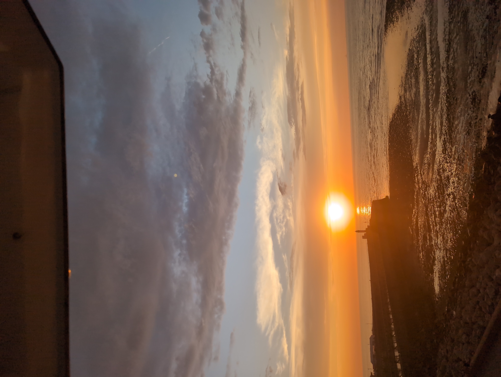 The image captures a sunset over the sea at Le Tréport, France. The sun, a bright orb of light, is low on the horizon, casting a warm, orange glow across the sky and water. The sky is a canvas of colors, transitioning from bright orange near the sun to hues of pink, yellow, and muted blue higher up, with scattered clouds creating texture and depth.
In the foreground, the rugged, rocky coastline is visible, with pools of water reflecting the vibrant sky. A man-made pier or walkway extends from the coast into the sea, lined with what appears to be railings. At the end of this structure, a lighthouse stands tall, its silhouette dark against the illuminated sky. Small figures, possibly people, can be seen on the pier near the lighthouse, adding a sense of scale to the scene. Further out at sea, the water is calm, with gentle ripples reflecting the sunset. The overall atmosphere is serene and picturesque, typical of a coastal sunset. The image is framed by a dark, horizontal element at the top, suggesting it might have been taken through a window or from a sheltered vantage point.
