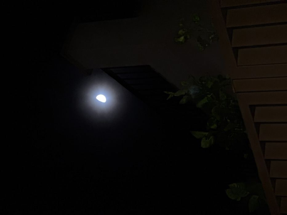 The image captures a nighttime outdoor scene, looking upwards towards a building structure. A bright, intensely glowing, half-moon shaped light source is prominent on the left side of the frame, emitting a strong halo that illuminates the immediate dark surroundings. To the right, architectural elements are visible, including a dark roof eave or overhang extending diagonally from the upper center, and a section of a building facade with horizontal siding on the far right. Dark green foliage, consisting of leaves and branches, is sparsely visible in the middle and upper right sections, appearing mostly silhouetted against the dark sky, with some subtle glints of light. The scene is static, devoid of any discernible activity or individuals. The time of day is clearly night, with no specific weather details discernable beyond the clear visibility of the light source. There is no visible text in the image. The location context is Beausoleil, France.