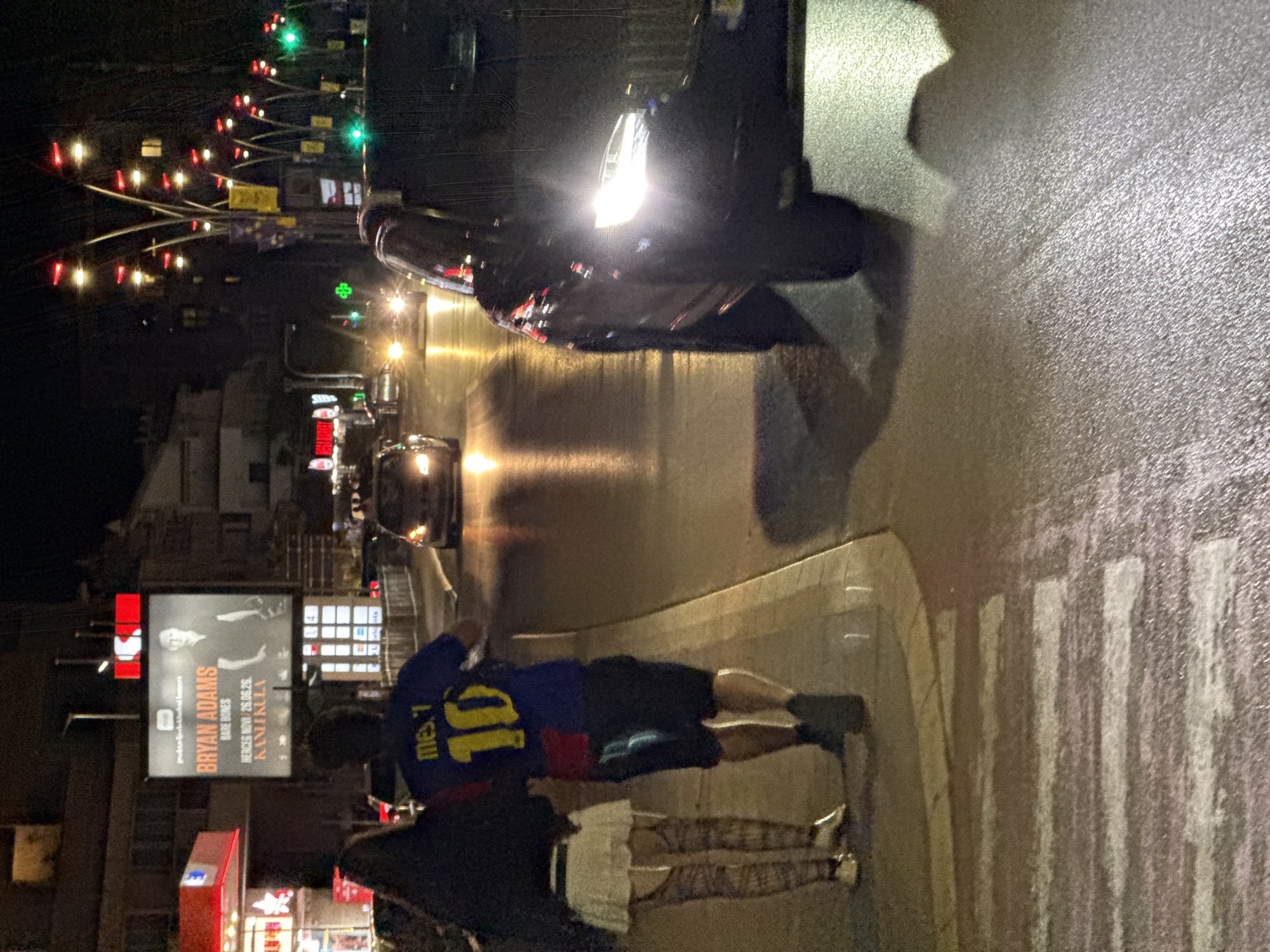 The image captures a nighttime scene on a street in Budva, Montenegro. In the foreground, a crosswalk is visible on the wet pavement, reflecting streetlights. To the left, two figures are walking away from the camera, with the person on the right wearing a blue and red soccer jersey with the name "MESSI" and the number "10" on the back. The person on the left is wearing a white skirt and patterned stockings. A dark-colored vehicle with its headlights on is in the right lane of the street, appearing to be in motion. Further down the street, other vehicles and buildings with illuminated signs are visible. A large billboard featuring Bryan Adams is prominently displayed on the left side of the street. The atmosphere suggests a typical urban environment at night, with artificial lighting creating reflections on the wet surfaces.