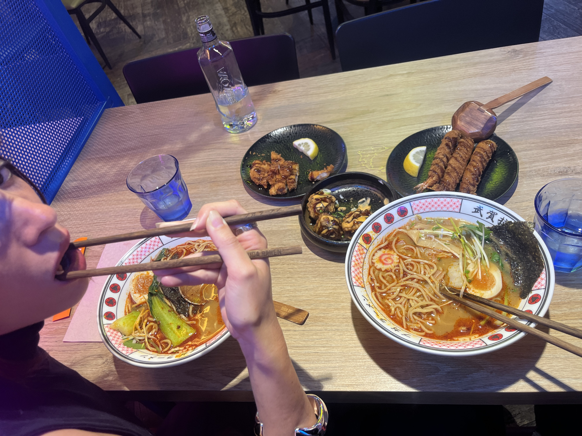A table setting shows two bowls of ramen, fried shrimp, and chicken karaage, with a person eating from one of the bowls using chopsticks. A glass of water and a bottle of water are present. The location is likely a restaurant in Barcelona, Spain.