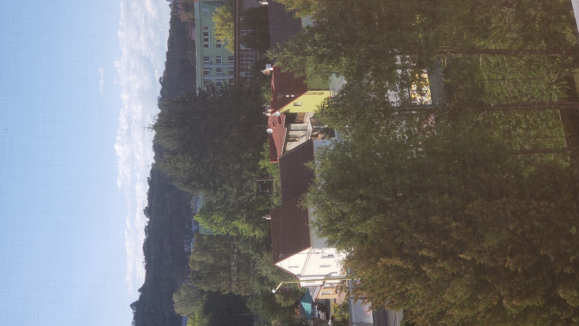 The image shows a residential area with several houses and a larger building in the background. The houses are of varying colors and styles, with pitched roofs and some featuring balconies. Trees and greenery are abundant throughout the scene, creating a suburban or semi-rural atmosphere. The sky is clear with some wispy clouds. A screen mesh is visible in the foreground, suggesting the image was taken from indoors through a window. The specific location in Prešov, Slovakia, cannot be determined from the visual information provided.