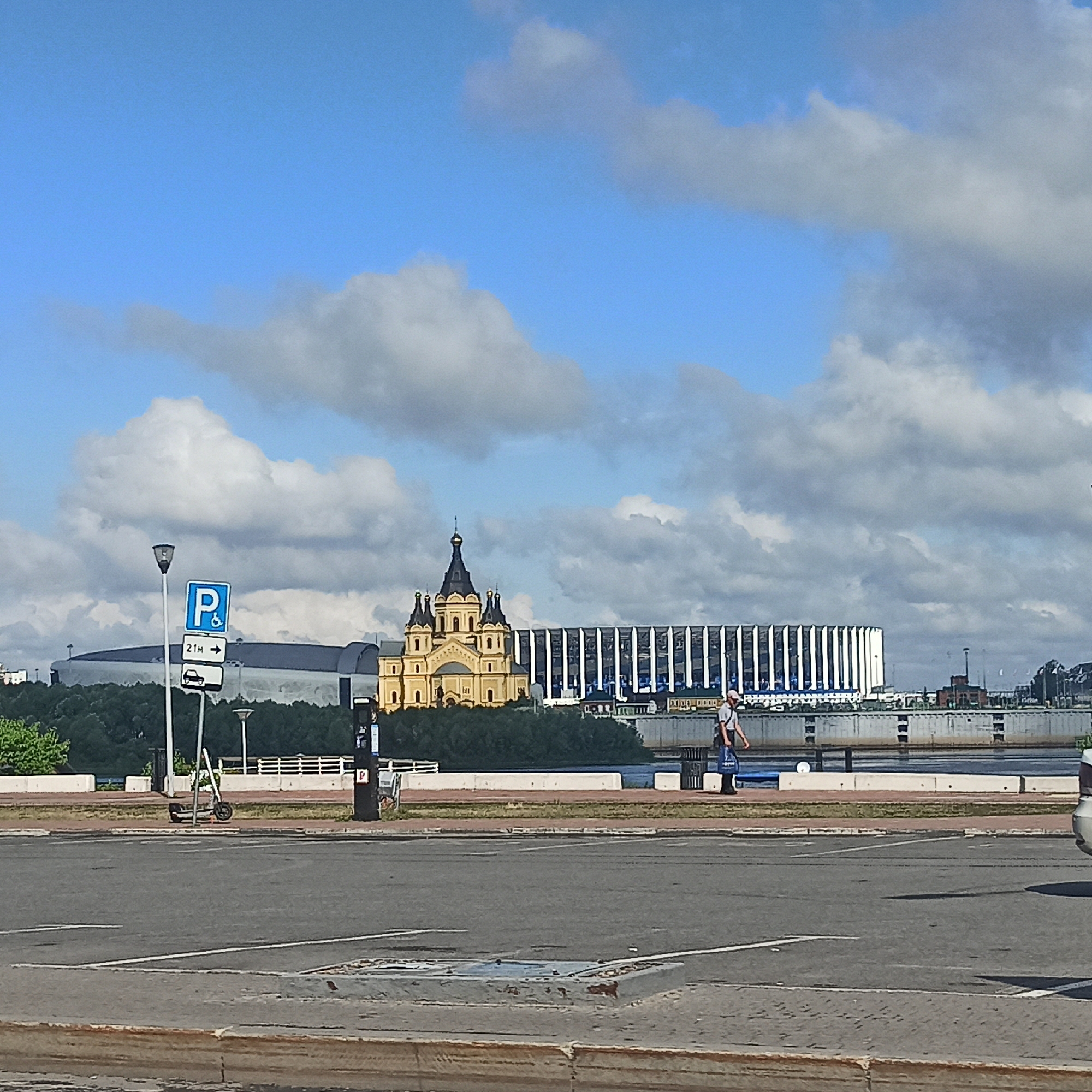 The image captures a daytime scene in Nizhny Novgorod, Russia, with a focus on the Volga River embankment and the cityscape. In the background, the modern stadium "Nizhny Novgorod Stadium" stands prominently, its white facade with vertical blue accents reflecting the sunlight. To the left of the stadium, a large, multi-domed yellow church with dark spires is visible, likely the Alexander Nevsky Cathedral, situated on a tree-lined embankment.
In the foreground, a paved road and sidewalk area is depicted. Several signs are present, including a blue and white parking sign with a wheelchair icon and another sign below it indicating "21m" with an arrow pointing right, and a car icon. A street lamp with a directional light fixture stands beside these signs. A black parking meter or ticket machine is also visible. On the sidewalk, a grey electric scooter is parked.
A pedestrian, a male wearing a light-colored t-shirt and dark pants, is walking from right to left across the embankment, carrying a blue bag. The Volga River is visible beyond the embankment, with a concrete barrier along its edge. The sky is blue with scattered white and grey clouds. The overall lighting suggests a bright, sunny day.