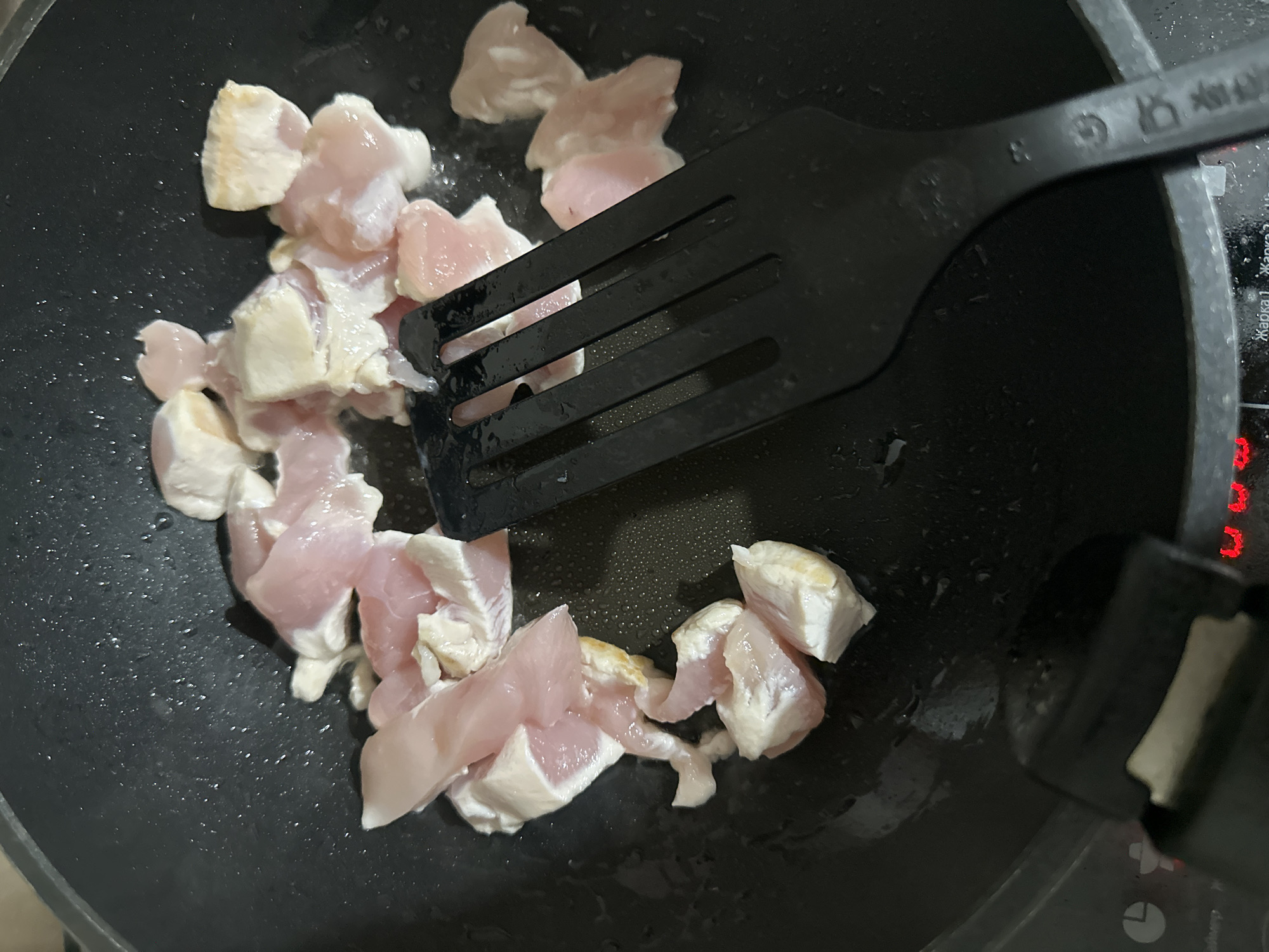 An overhead close-up shot shows pieces of raw or lightly cooked chicken being prepared in a black non-stick pan. The chicken, cut into cubes or strips, displays a pinkish color with some white fat and connective tissue, suggesting it's in the early stages of cooking. A black slotted spatula is positioned among the chicken pieces, indicating active stirring or flipping. Small droplets of oil or moisture are visible on the pan's surface.
The scene takes place in a kitchen, with part of an electric or induction stovetop visible on the right. This stovetop features a red digital display, showing a segment that resembles the letter "n," along with circular icons, one of which appears to be a timer symbol. Crucially, Cyrillic text is visible on the stovetop, reading "Жарка 1" and "Жарка 2," which translates to "Frying 1" and "Frying 2." These details provide a strong location cue, consistent with the given context of Perm, Russia.
The black spatula has faint markings on its handle, including what looks like the number "8" and two small symbols: one resembling a thermometer, implying heat resistance, and another that looks like a glass and fork, commonly used to denote food-safe materials. The image captures a routine cooking activity, likely preparing a meal. The time of day is not specified, but the indoor setting is illuminated by artificial light. No people or discernible emotions are visible.