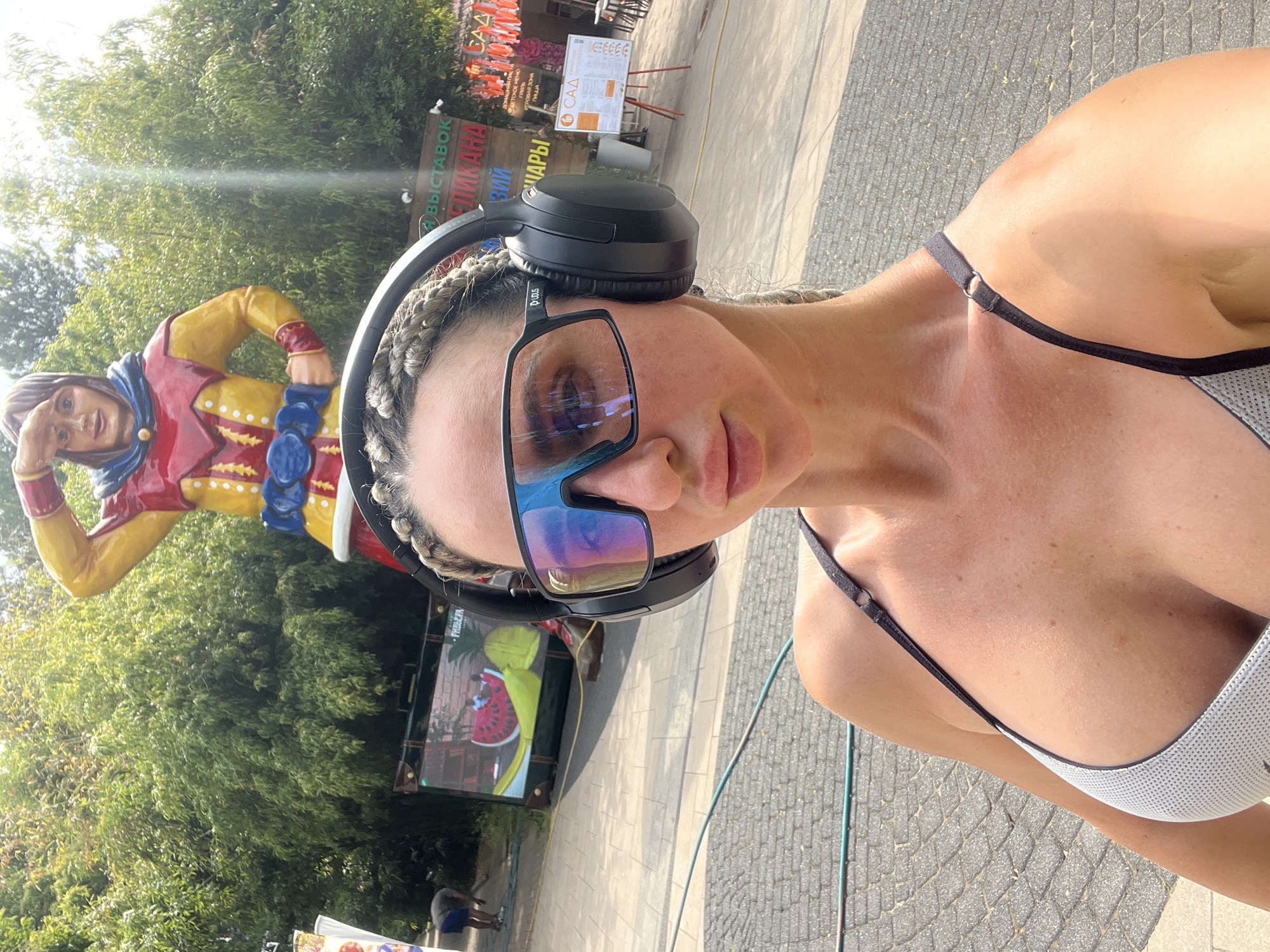 A woman is taking a selfie outdoors. She is wearing black headphones and sunglasses, and a grey and black sports bra. Her hair is braided. In the background, there is a colorful statue of a man in a jester-like outfit. Behind him, there are trees and a sign that says "А ВЫСТАВОК" and other Russian text, likely indicating an exhibition or a park. The ground is paved with cobblestones, and sunlight suggests it is daytime. The setting appears to be a public area in Sochi, Russia, possibly a park or an outdoor exhibition. There are also some distant figures visible, suggesting other people are present. The image is oriented vertically.