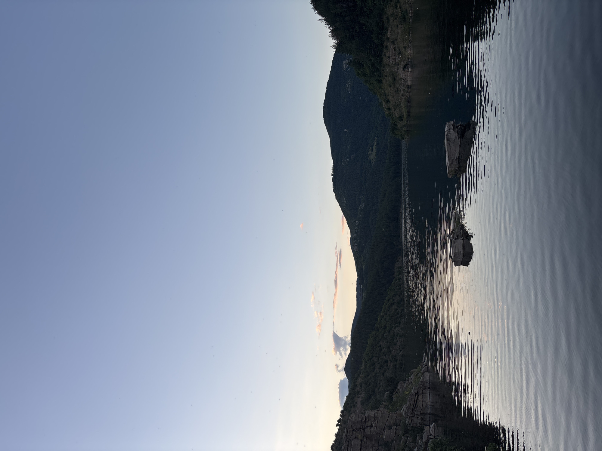 This is a vertical, eye-level, long shot of the dam at Suhovo, Bulgaria, taken during twilight. The sky is a pale, clear blue, fading to a lighter hue near the horizon where a thin strip of pinkish-orange clouds is visible. Two large, dark, rugged mountains rise from the far side of a calm, dark body of water, their slopes covered with dense, dark green pine trees. The water reflects the sky and the surrounding landscape, creating rippling patterns. In the middle of the water, two large, rectangular rocks protrude from the surface. On the left side of the image, the rocky, steep bank of the dam is visible, with sparse vegetation growing on its surface. The overall mood is serene and peaceful.