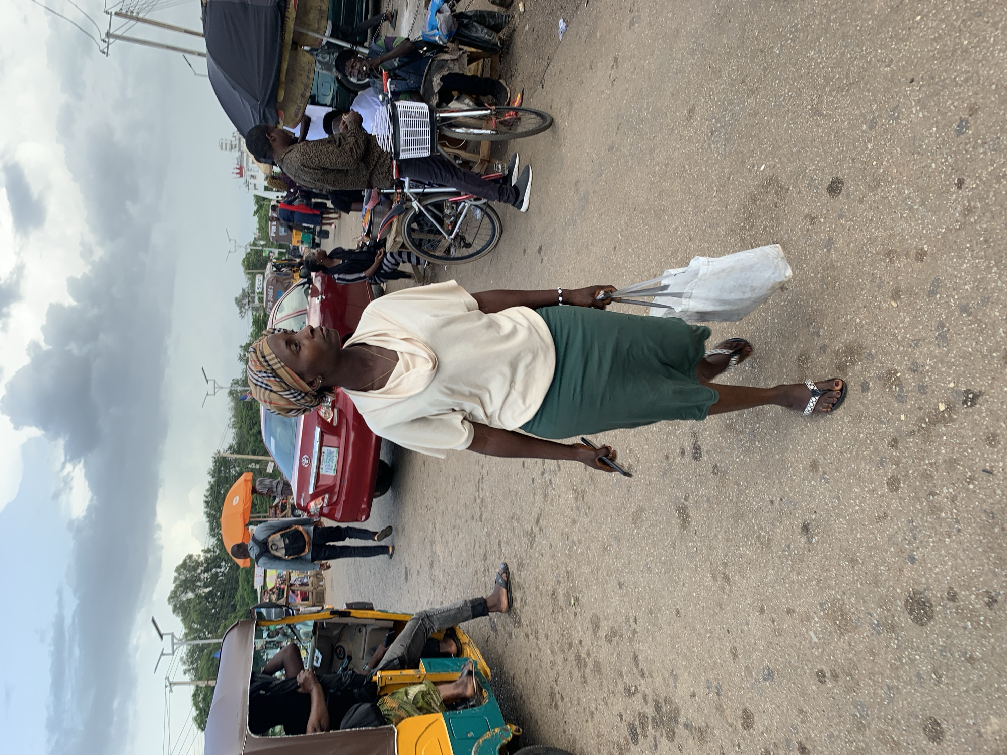A woman walks across a dusty road in Jos, Nigeria, carrying a white bag and a dark phone. She wears a beige cowl-neck top, a green skirt, and patterned flip-flops. Her head is wrapped in a patterned scarf. In the background, a red car with a Nigerian license plate is parked. To the left, an auto-rickshaw, commonly known as a "Keke," is visible with passengers. Further back, people are seen near market stalls and a bicycle. The sky is overcast with scattered clouds. The scene appears to be a busy street market area.
