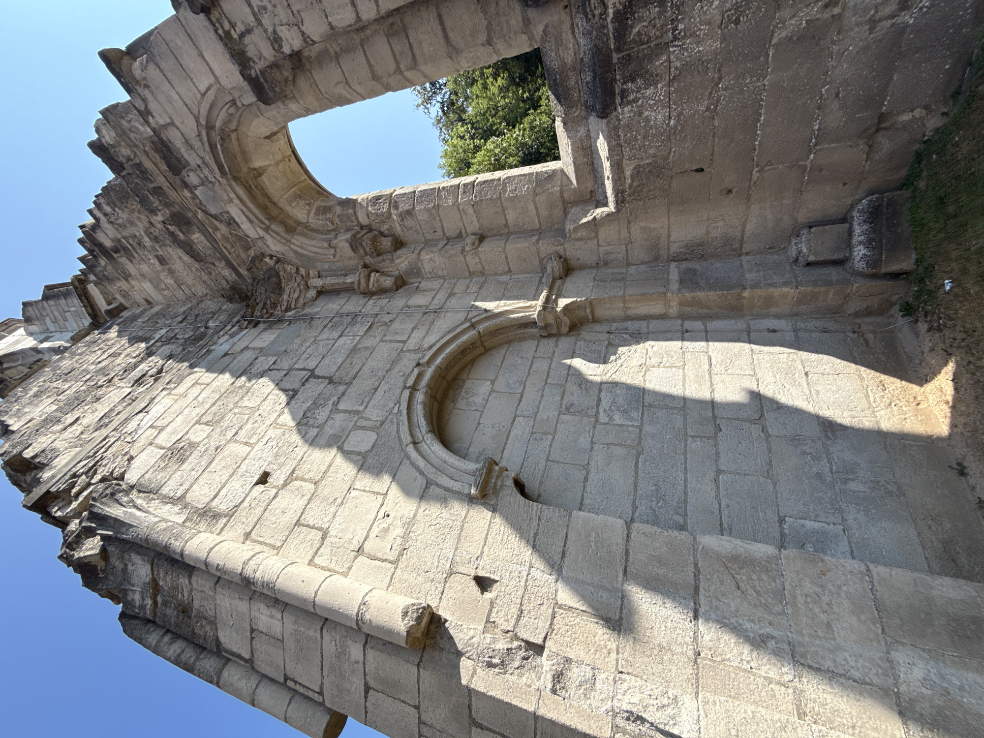 The image shows a close-up, low-angle view of the ruins of the Walkenried Abbey in Walkenried, Germany. The perspective emphasizes the weathered stone construction of a partially standing wall, with large, roughly cut blocks forming the structure. A significant portion of the wall is missing, exposing the clear blue sky and some green foliage in the background through two arched openings. One opening is a large, grand arch, while a smaller, more complete arched niche is visible on the left side of the wall. A distinct shadow, cast by an unseen object, stretches across the stone surface, highlighting the texture and contours of the stonework. The light is bright, suggesting a sunny day. The ruins appear to be ancient, with signs of erosion and age visible on the stone. There are no people or animals present in the image. The ground beneath the wall shows sparse dry grass and some debris. The overall impression is one of historical decay and architectural grandeur.