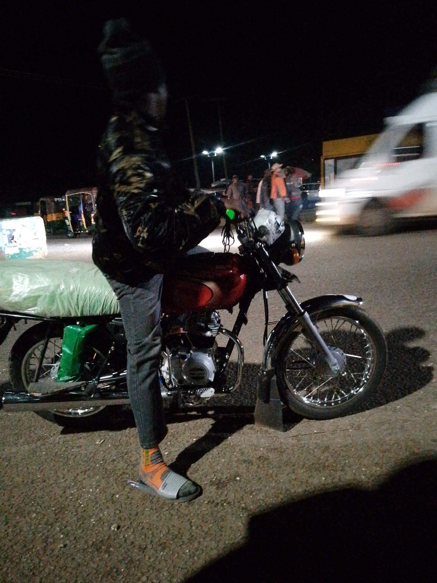A low-angle, full-body shot captures a person standing next to a red motorcycle at night. The person is wearing a dark, patterned jacket, gray pants, orange and gray socks, and sandals. The motorcycle is parked on a gravelly surface, with its rear covered by a green tarp. In the background, several auto-rickshaws and a white vehicle are visible, along with streetlights and the blurred outline of buildings. The overall scene suggests a street setting in Bukuru, Nigeria, possibly a transport hub or marketplace at night.