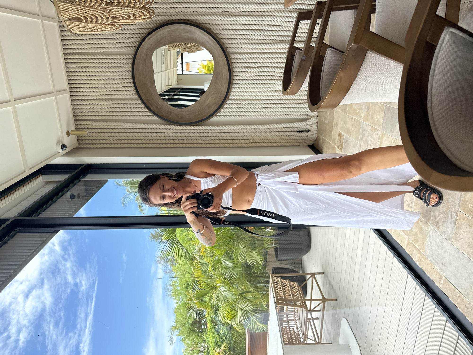 A smiling woman is captured in a mirror selfie, standing in a contemporary indoor-outdoor setting. She is wearing a white two-piece outfit that reveals her midriff and has a high slit, paired with black sandals. Her dark hair falls over her shoulders, and her left arm displays tattoos and bracelets. She holds a black Sony α7 camera, with "SONY" and "α 7" clearly visible on its strap, aimed towards her reflection.
The interior space features a unique design with a wall or ceiling covered in thick, horizontal woven ropes. A round wooden-framed mirror hangs on this rope surface, reflecting parts of the room, including wooden chairs with light-colored cushions and more rope texture. A small, white security camera and a gold-colored fixture are mounted on a cream-colored panel in the upper section of the interior. The floor is made of light-colored, patterned stone tiles.
The woman is positioned in front of a large glass window or partition, which vividly reflects the outdoor environment. The reflection shows a vibrant tropical scene with lush green palm trees and other dense foliage under a bright blue sky with scattered white clouds, suggesting clear daytime weather. An outdoor deck with light-colored wooden planks is visible, along with what appears to be a wicker table and chairs. The scene evokes a sense of relaxation and leisure in a warm, resort-like location, consistent with the provided context of Ko Pha Ngan, Thailand.