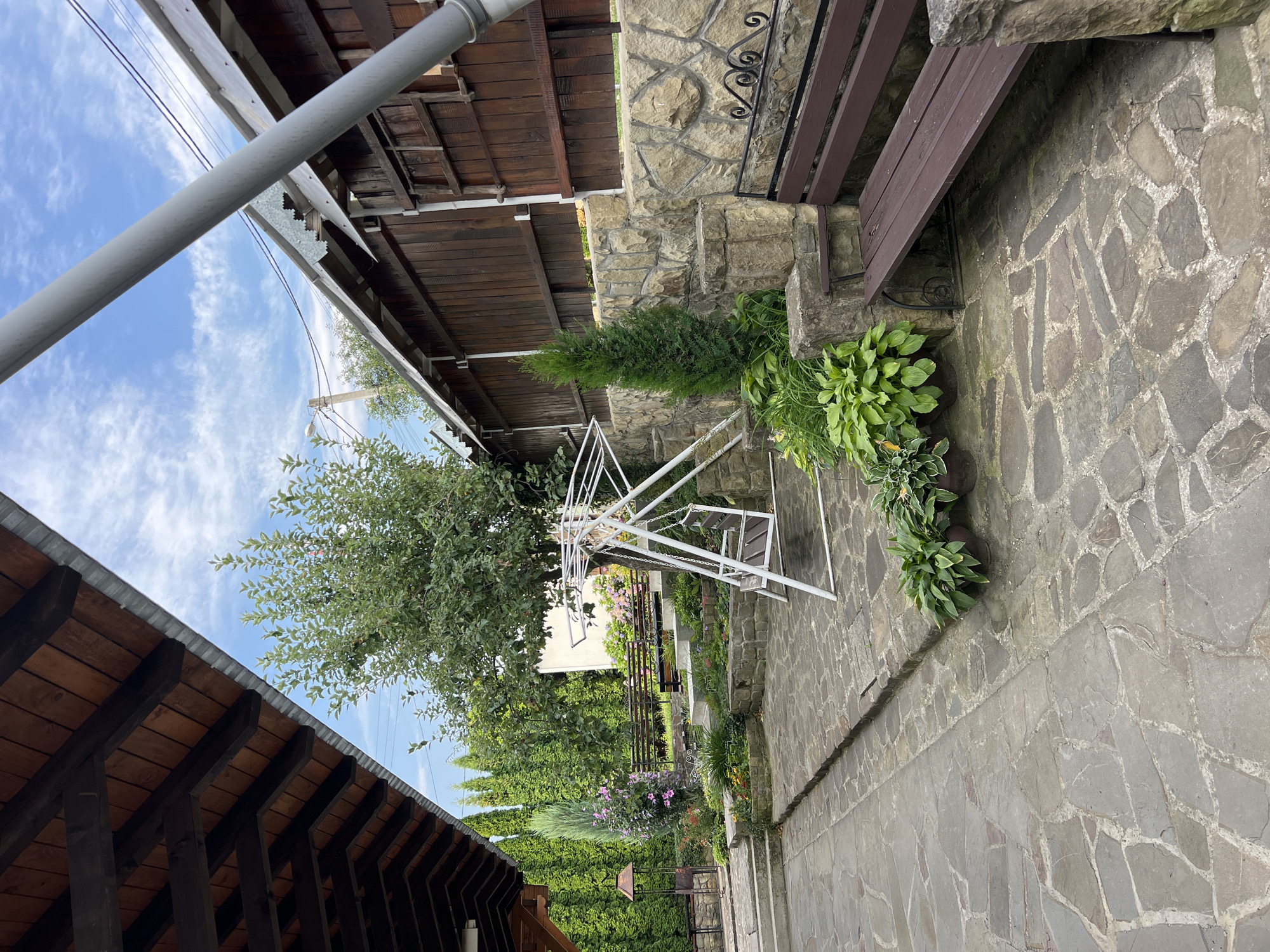 The image depicts an outdoor courtyard with a rustic aesthetic, likely a part of a guesthouse or private residence in Yaremche, Ukraine. The ground is paved with irregular flagstones. To the left, a wooden roof structure with exposed beams extends over a portion of the courtyard, with a metal gutter system visible. Further back, a tiered garden area is visible, featuring a white metal swing set, various potted plants including hostas, a small evergreen tree, and flowering plants with pink and purple blossoms. Stone retaining walls are incorporated into the garden's design, supporting the different levels. A dark wooden bench with metal scrollwork accents is positioned to the right, adjacent to the stone wall. The background consists of a wooden fence, more greenery, and a cloudy blue sky. The scene is devoid of people, and no specific actions or interactions are occurring. The overall impression is one of a well-maintained, tranquil outdoor space.