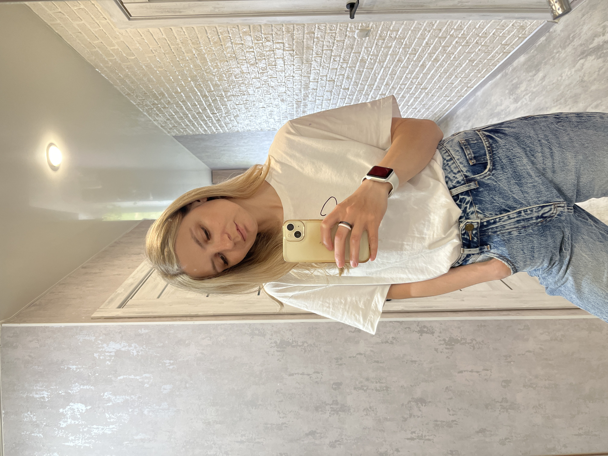A person is taking a selfie in a bathroom, in an indoor setting. The person appears to be a young, light-skinned woman with blonde hair. She is wearing a white t-shirt and blue jeans, and a white smartwatch on her left wrist. She is holding a gold smartphone, presumably to take the picture. The bathroom has a gray and white color scheme with textured walls and white doors. There is a light fixture visible on the wall and a patterned ceiling in the background. Given the prompt, the location is likely in Chaikovsky, Russia.