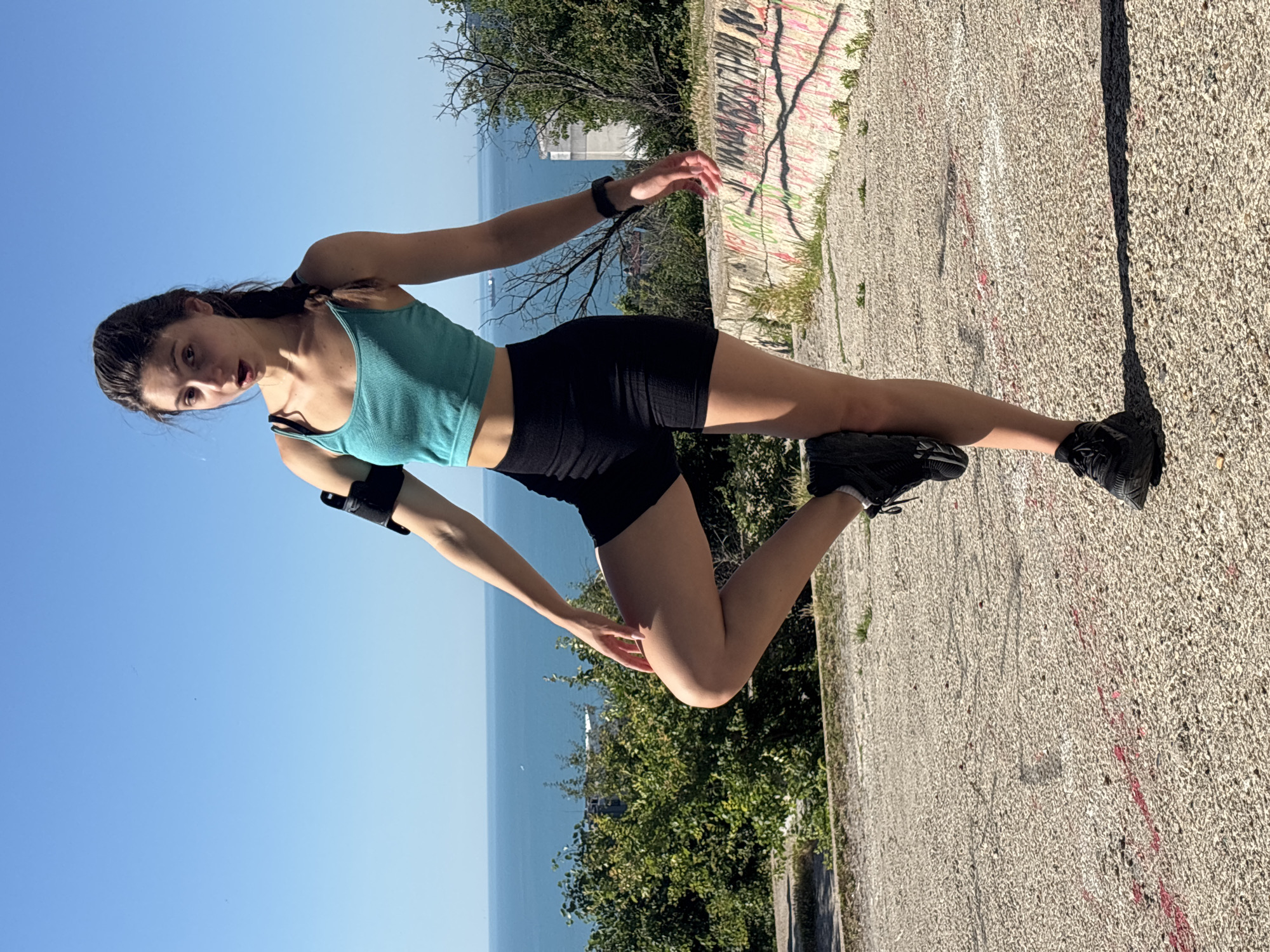 A young woman in athletic attire stands on a concrete surface with a body of water and a cityscape visible in the background under a clear blue sky. The woman is wearing a teal sports bra, black athletic shorts, and black sneakers. She is holding her right leg up behind her, balancing on her left leg in a pose that appears to be for exercise or a photograph. She has a phone armband on her left bicep and a fitness tracker on her left wrist. The concrete surface is weathered and appears to have some red and white markings. A low, graffiti-covered wall is visible behind her to the right, with lush green bushes and trees extending towards the water. The presence of the sea and distinct architectural styles in the background suggest the location is likely Varna, Bulgaria, specifically an elevated area overlooking the Black Sea, possibly near the Sea Garden or a similar coastal park. The lighting indicates it is daytime, with the sun casting shadows.