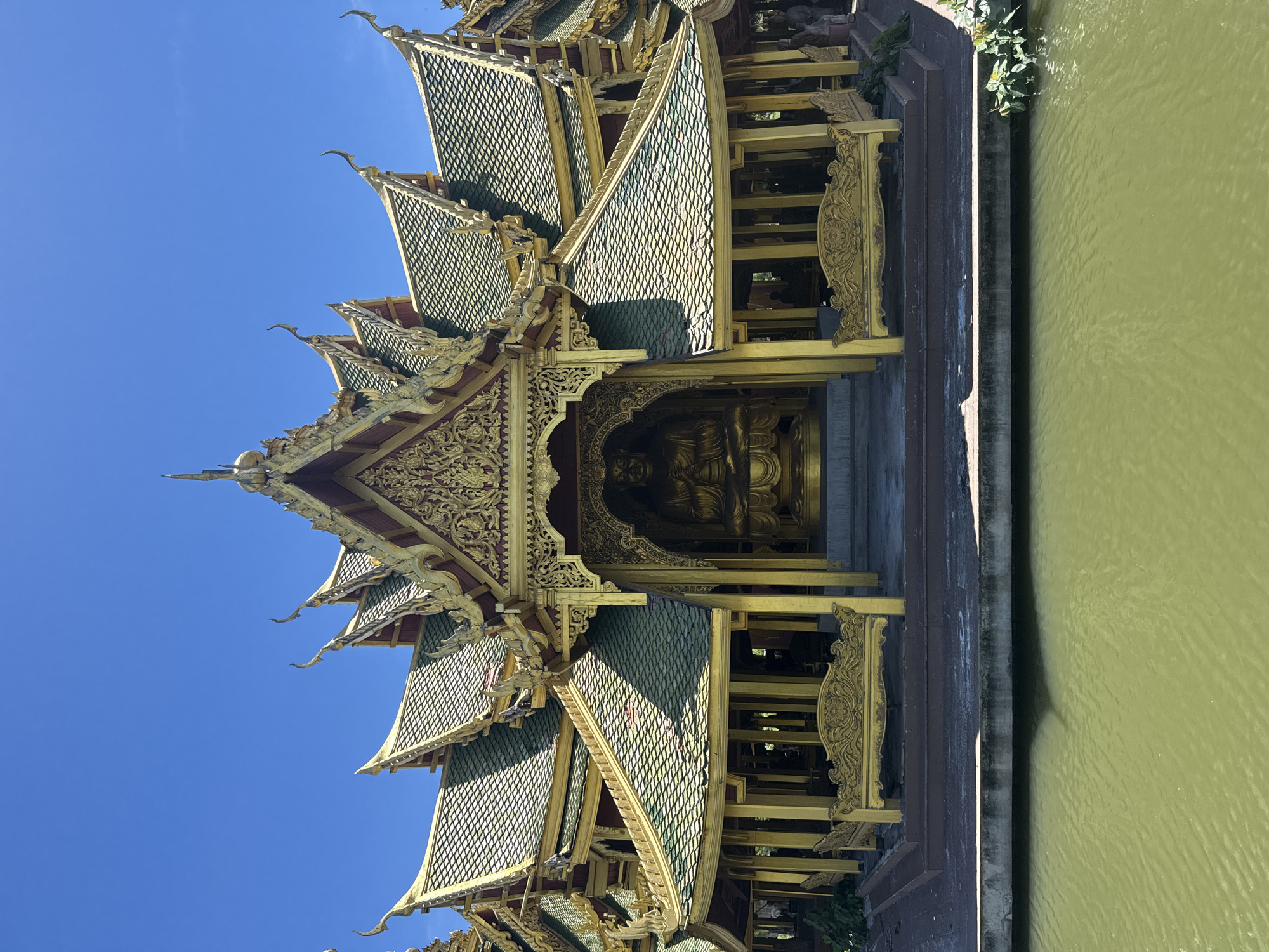 The image shows a detailed view of a traditional Thai pavilion located within the Ancient City (Muang Boran) in the Bang Pu Subdistrict Municipality, Thailand. The pavilion has a tiered roof design with green tiles, ornate gold trim, and intricate carvings. A golden Buddha statue is visible inside the pavilion. The pavilion is situated next to a body of water. The sky is clear and blue.