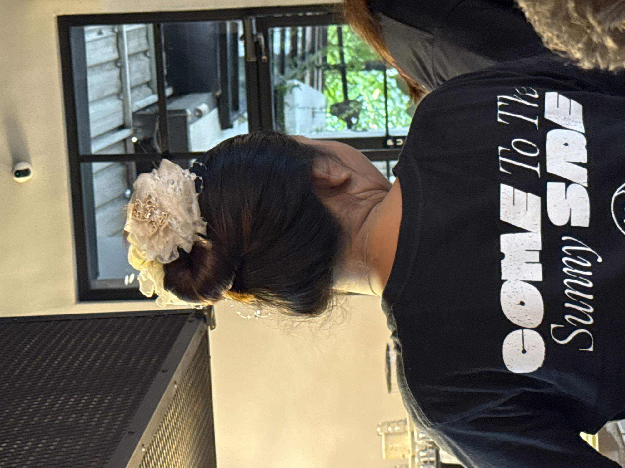 A person wearing a black t-shirt with the words "Come To The Sunny Side" is standing in what appears to be an indoor setting. The person has dark hair styled in a bun adorned with a white floral accessory. A black speaker is located in the foreground to the left. In the background, there is a window showing an exterior view with greenery. Based on the context of the location and surrounding scenery, the image may be from North Kuta, Indonesia.