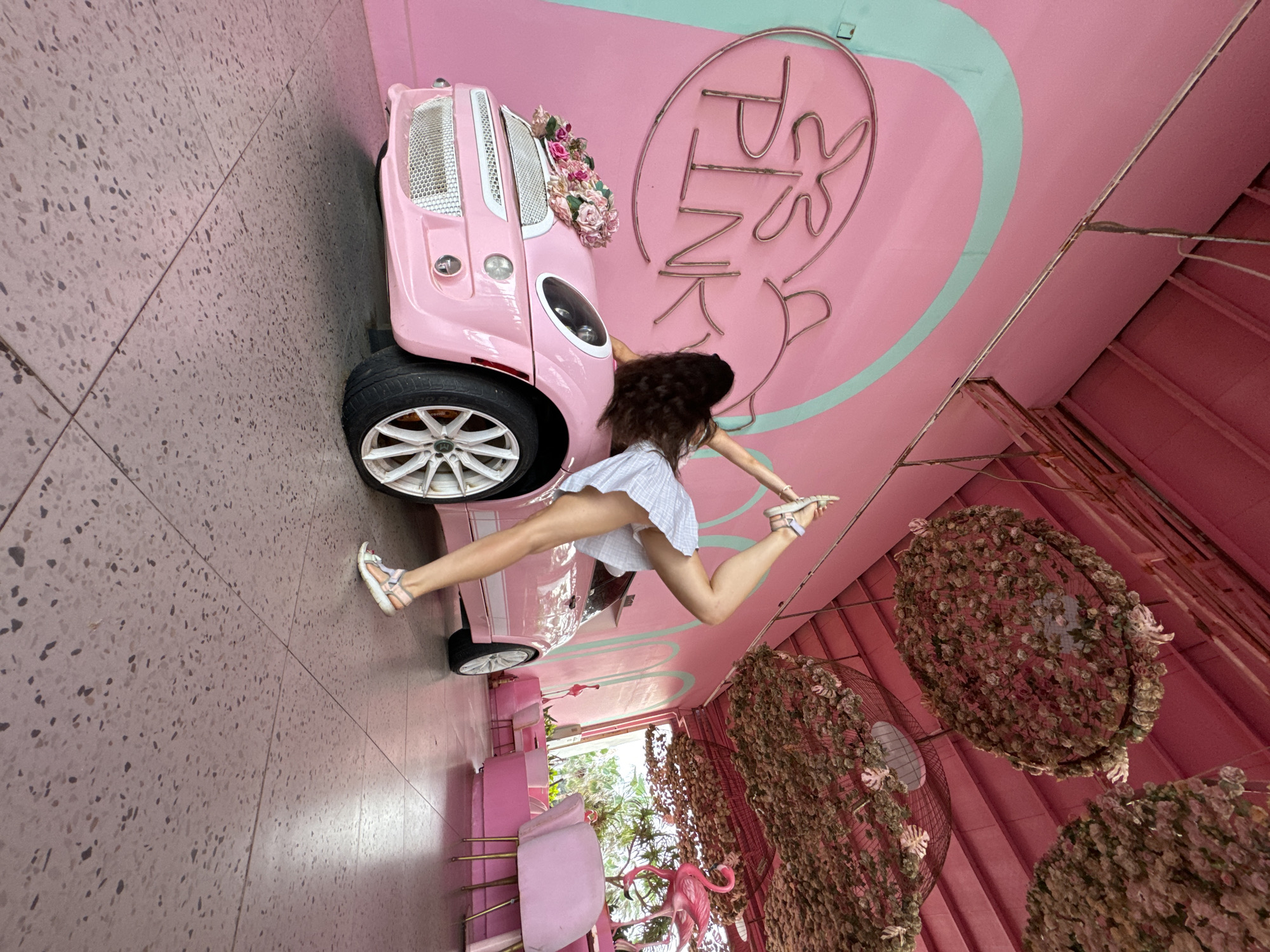 A woman performs a yoga pose next to a pink Mini Cooper in a pink room with a flamingo statue and a neon sign that says "PINKY". The ceiling is adorned with hanging floral decorations. The floor is a patterned pink terrazzo.