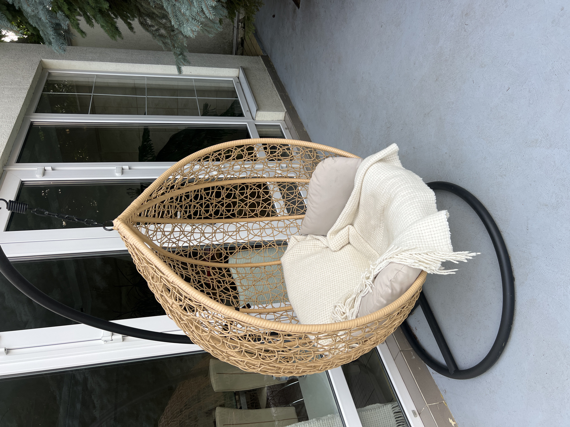 An outdoor wicker hanging chair with a tan cushion and a cream-colored knitted throw blanket is positioned on a grey concrete patio next to a white-framed glass door. A portion of a dark green evergreen tree is visible on the right side of the image. The scene appears to be a residential outdoor space.