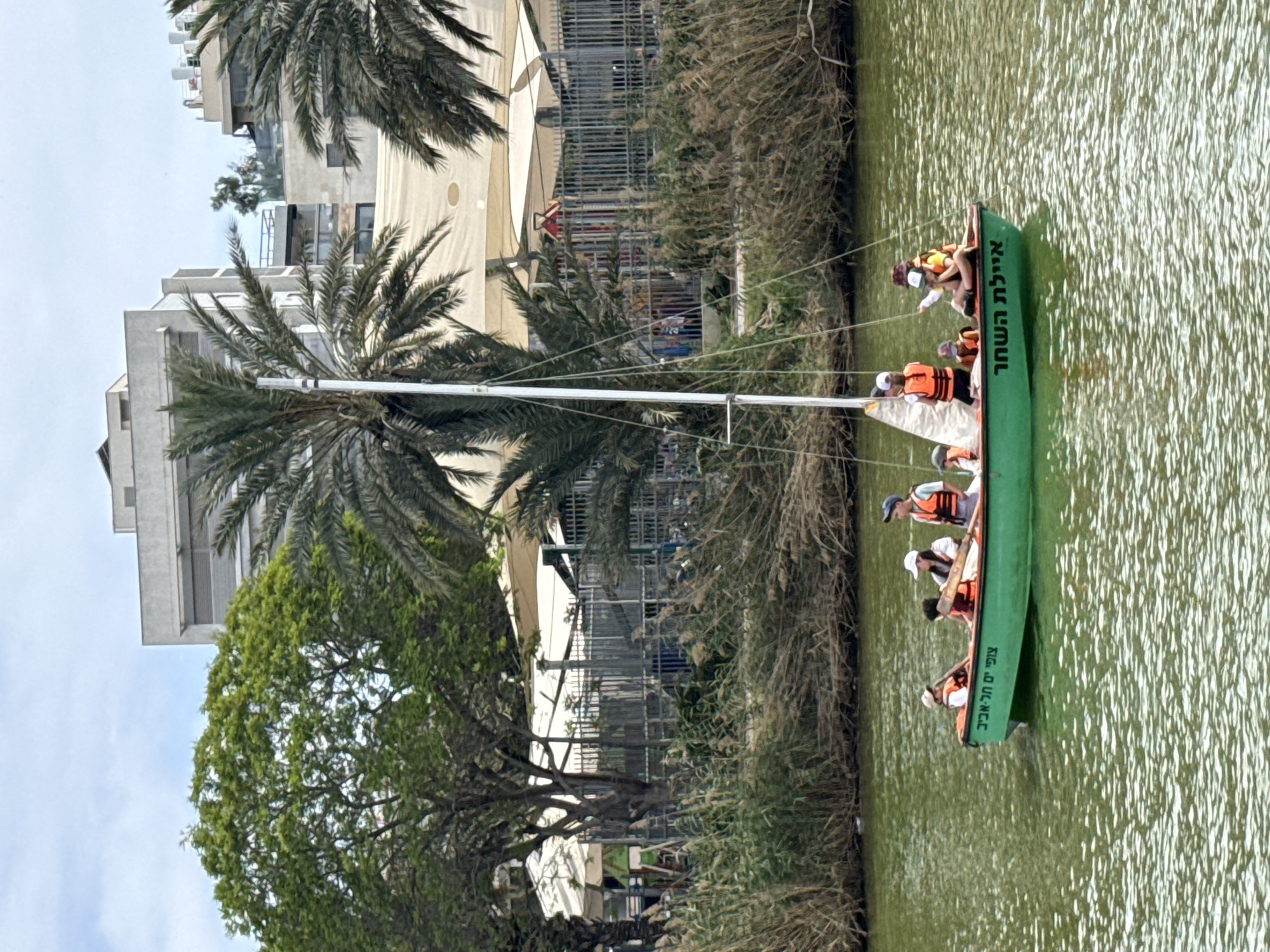 A boat with people wearing orange life jackets is sailing on the Yarkon River in Tel Aviv, Israel. The green boat has Hebrew writing on its side, naming the tour location. Vegetation lines the riverbank, and buildings and palm trees are in the background.