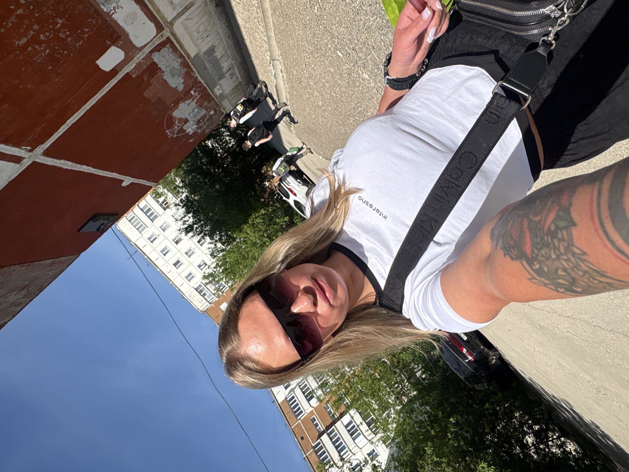 The image shows a woman taking a selfie outdoors on a sunny day. She is wearing sunglasses, a white t-shirt with black trim, and a black purse with a Calvin Klein strap. Her left arm has a tattoo. The background shows buildings, trees, parked cars, and pedestrians. Based on the architectural style and streetscape elements, this image was taken in Perm, Russia.