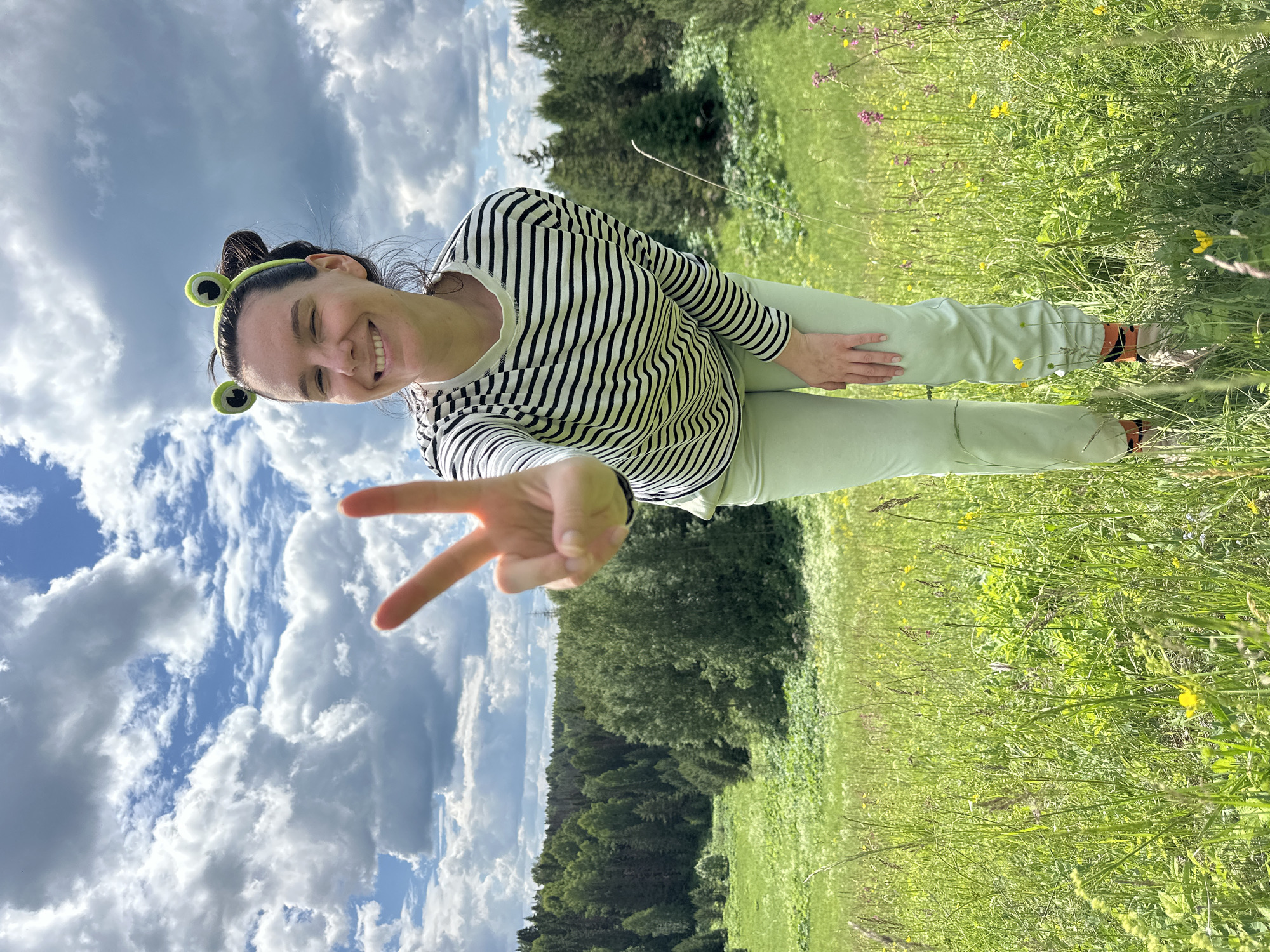 A young adult female, centered in the frame and leaning slightly forward, smiles broadly with teeth showing and winks with her left eye. She wears a bright green headband with two prominent, round frog-eye attachments. Her attire consists of a long-sleeved shirt with thin horizontal black and white stripes, light mint green trousers, and partially visible light-colored footwear with orange socks featuring dark patterns. Her right hand is extended towards the camera, forming a "peace" sign (V-sign) with her index and middle fingers. The scene is an open, sunlit grassy meadow on a gentle slope, filled with green foliage and scattered yellow and magenta wildflowers. In the midground and background, a dense mixed forest of various tree types covers rolling hills. The sky above is bright blue with numerous large, fluffy white and gray cumulus clouds. The location is in the vicinity of Альняш, Russia.