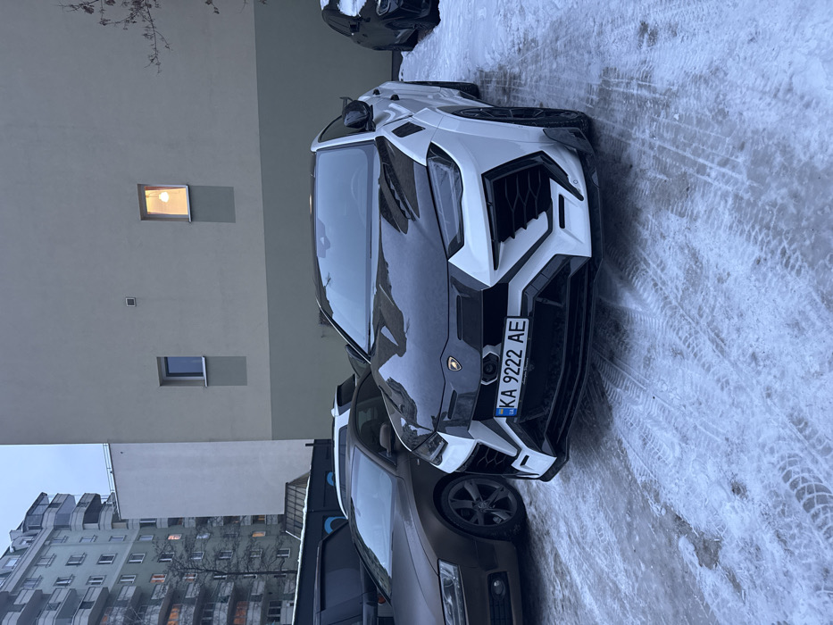 A black and white Lamborghini Urus is parked on a snowy surface next to a brown car. The Urus has a Ukrainian license plate that reads "KA 9222 AE." In the background, there is a building with lit windows and another taller building with multiple windows. The image is taken from a slightly elevated perspective, and the buildings appear to be urban or suburban.