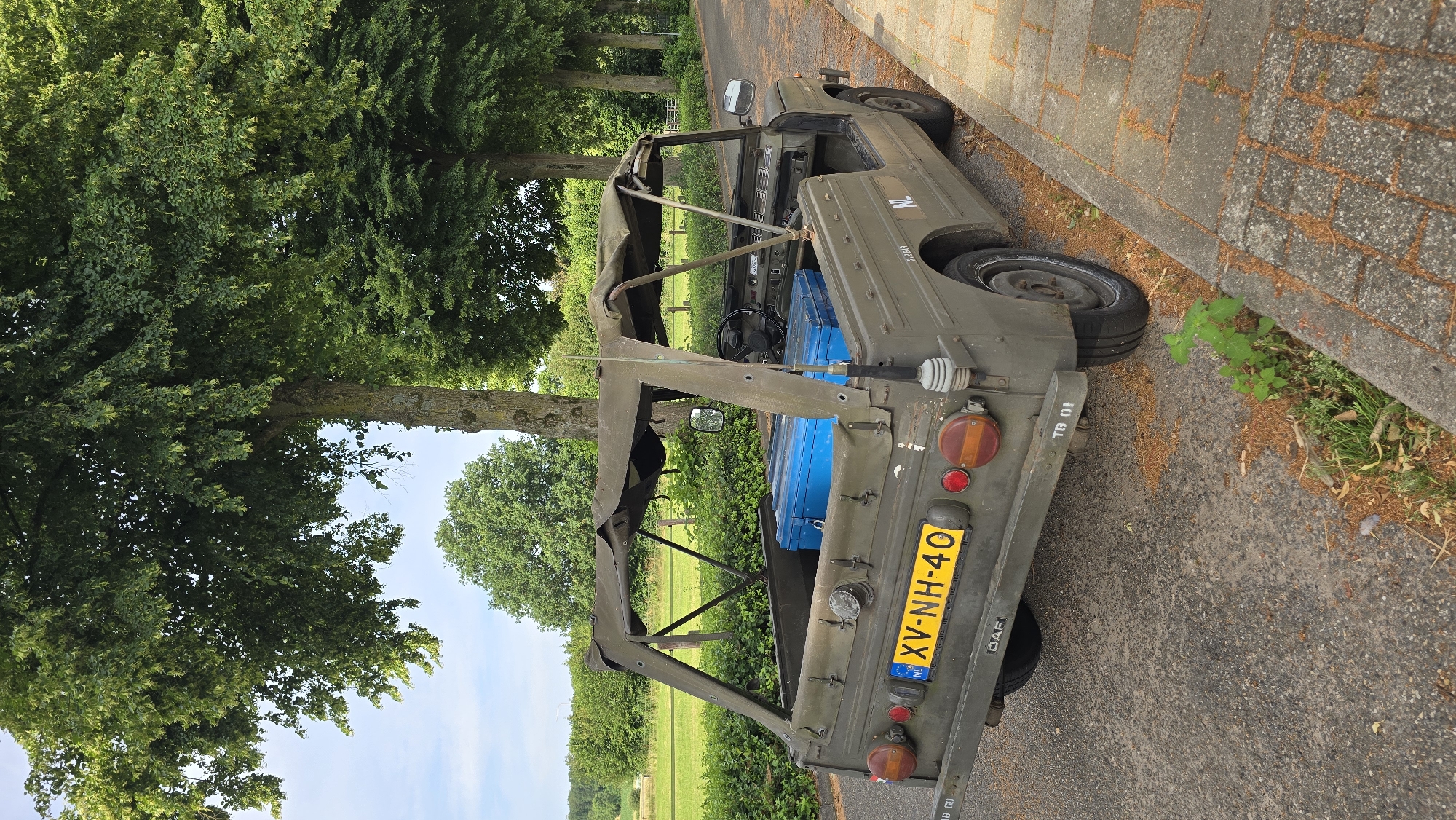 A military-style vehicle, possibly a DAF, is parked on the side of a paved road in Zeddam, Netherlands. The vehicle is olive green and has a canvas top. Two bright blue storage containers are visible in the open cargo area of the vehicle. The license plate reads "XV-NH-40". The road is lined with trees and green grass. The sky is blue with some clouds. The scene appears to be a sunny day.