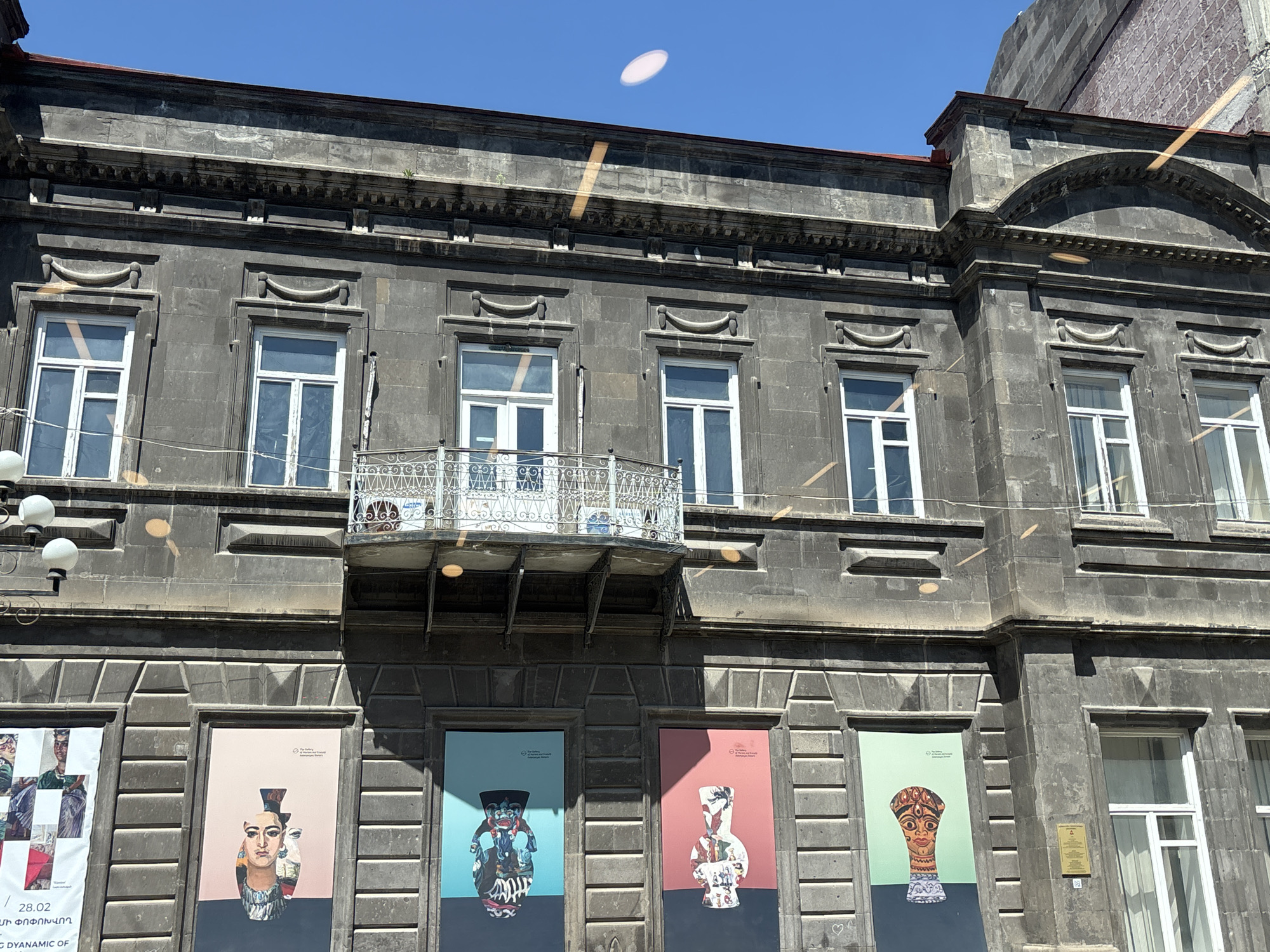 The image shows a grey stone building in Gyumri, Armenia. The building has two stories visible in the image, with five evenly spaced windows on each floor. The upper windows are rectangular with white frames, with decorative arched elements above each. The central window on the upper floor has a white metal balcony. The lower windows are similarly spaced and framed, but they are partially obscured by large posters featuring images of artifacts. A street lamp with three spherical lights is visible on the left of the image. The sky above the building is clear blue with a lens flare.