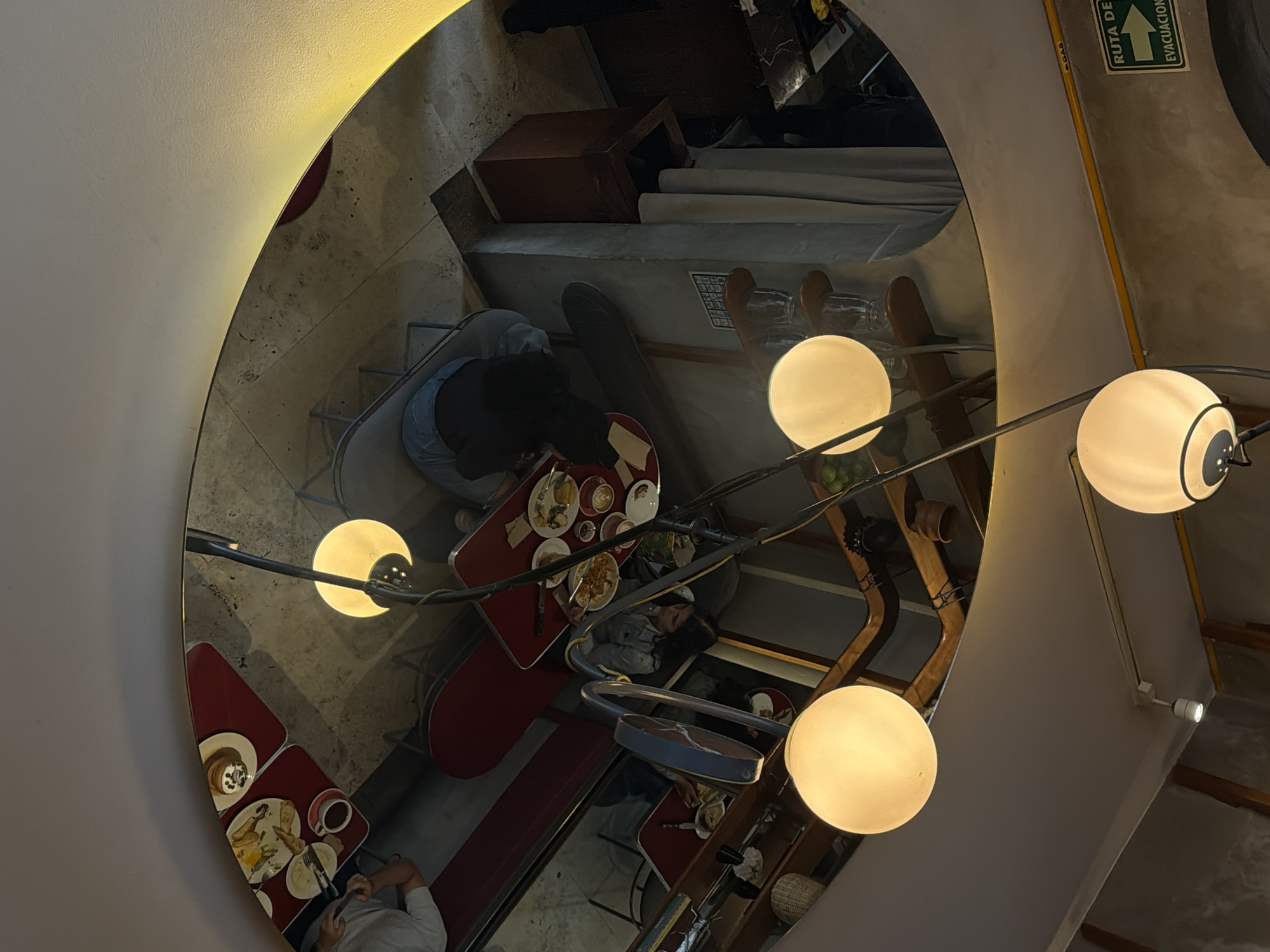 The image shows a distorted reflection of a restaurant in Mexico City, Mexico, as viewed through a concave mirror or other curved reflective surface. The mirror is mounted on a wall, which is visible in the upper and lower corners.
In the reflection, several red-topped tables are arranged with chairs around them. Diners are seated at the tables, eating and conversing. Some tables have plates with food on them, along with cups and cutlery.
A multi-bulb light fixture hangs in the foreground, with three visible spherical light bulbs. The fixture's arm crosses through the reflection, adding to the distortion. The restaurant's interior includes a bar area, with shelves and various objects. A sign that appears to be an evacuation route is visible on the upper-right side of the mirror's frame.