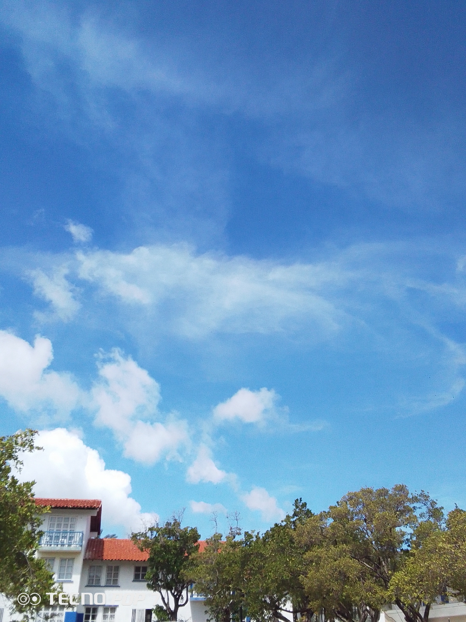 The image shows a bright blue sky with scattered cumulus clouds, partially obscuring a white, two-story building with a red tile roof. The building has a balcony with blue railings. Green trees partially surround the building. The image was taken outdoors during daylight. The location is likely Pedro González, Venezuela, as specified. The "Tecno Pop" watermark suggests the image was taken with a Tecno Pop smartphone.