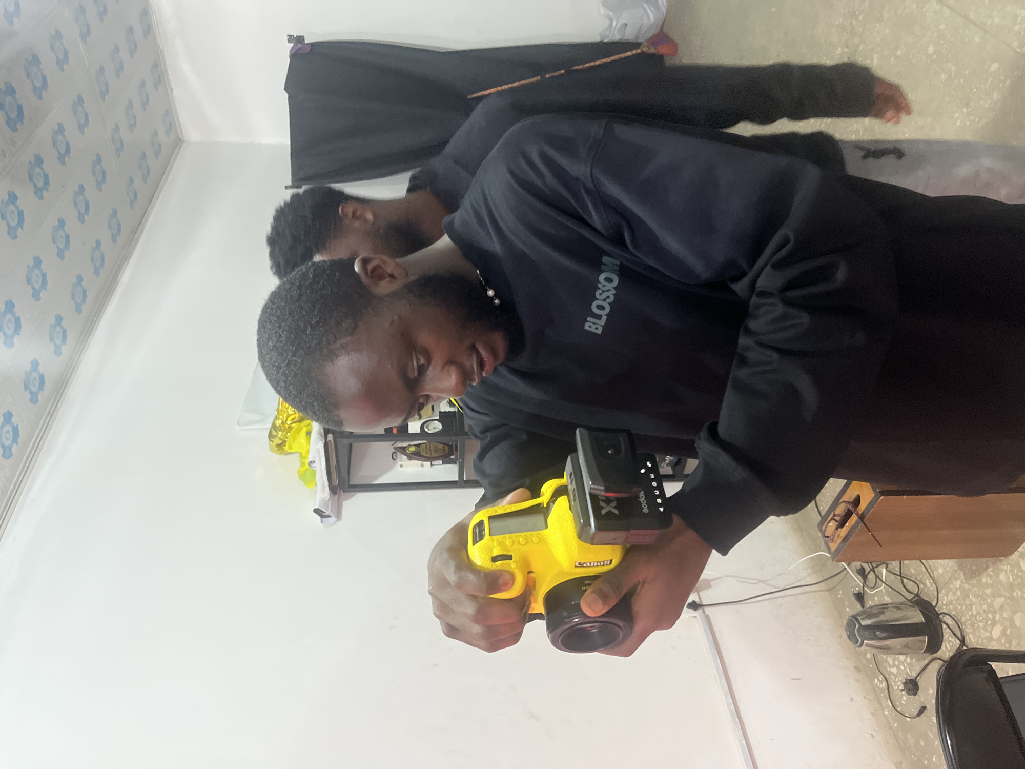 In Gura Top, Nigeria, a young Black man wearing a black crewneck sweatshirt with "BLOSSOM" printed on it, is holding a bright yellow camera with a black lens and a small black attachment on the front. He is looking down at the camera with a focused expression. Behind him, another Black man, also wearing black clothing, stands partially obscured. The environment appears to be an indoor space with white walls and a ceiling decorated with a repeating blue floral pattern. To the left, an electric kettle is plugged into a wall socket, and various cables are visible on the floor. A wooden object, possibly a speaker or shelf, is also present. The overall scene suggests a casual, possibly creative setting where photography is taking place.