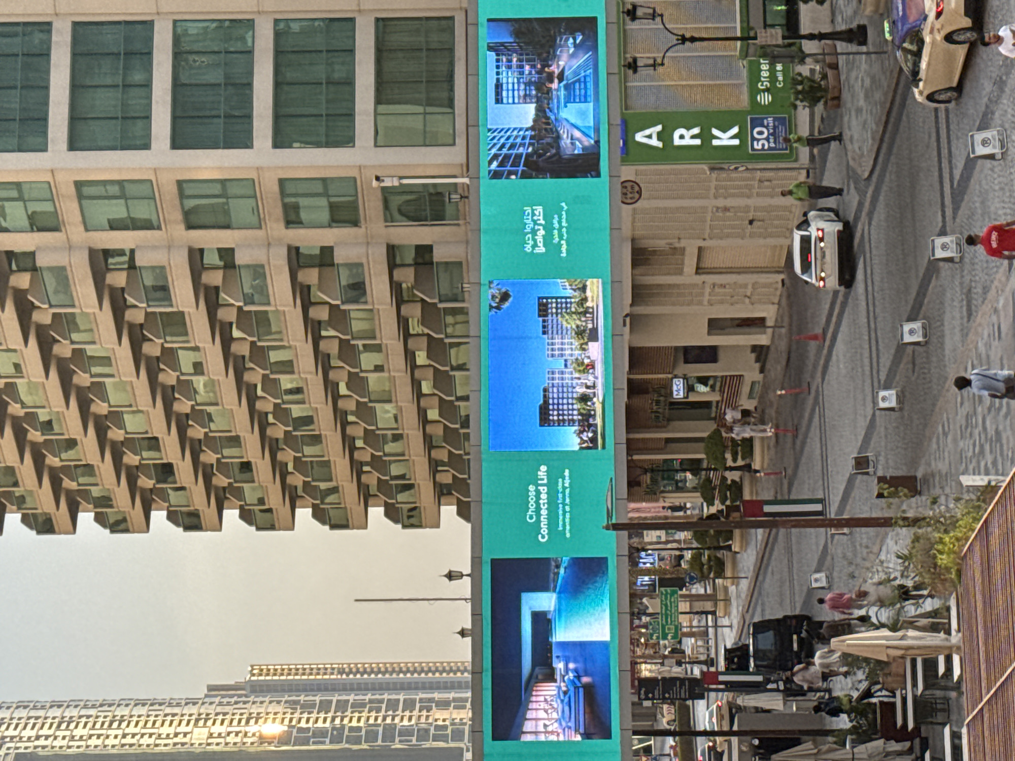 The image depicts a street scene in Dubai, United Arab Emirates, with a large digital billboard advertising real estate. The billboard displays images of modern architecture and swimming pools, along with text in both English and Arabic. The English text reads "Choose Connected Life" and "Immersive first-class amenities at Jenna, Aljada." The Arabic text appears to translate to similar messaging.
In the foreground, the street is paved with cobblestones and lined with shops and restaurants. Several people are walking on the street, some dressed in casual attire, while others appear to be employees or residents. A black SUV and a white sedan are parked on the street, and a yellow taxi cab is visible in the distance. The buildings in the background are tall and modern, characteristic of Dubai's cityscape. The overall atmosphere is that of a bustling urban environment.