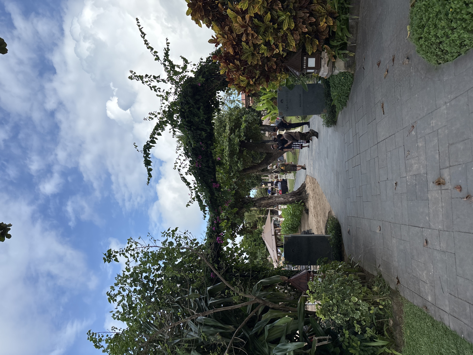 The photograph depicts a paved pathway flanked by dense vegetation, possibly a garden or landscaped area, in Kuta Selatan, Indonesia. A large sign that reads "PARADISUS" and "WELCOME" is partially visible on the right side of the path. Several people are walking along the pathway towards the center of the image. The vegetation includes various trees and shrubs, some of which are trained to form a canopy over the pathway. The sky is visible above the trees, featuring a mix of blue sky and white clouds. The photo appears to be taken at ground level with a slight upward tilt. The overall impression is that of a tranquil, well-maintained outdoor space.