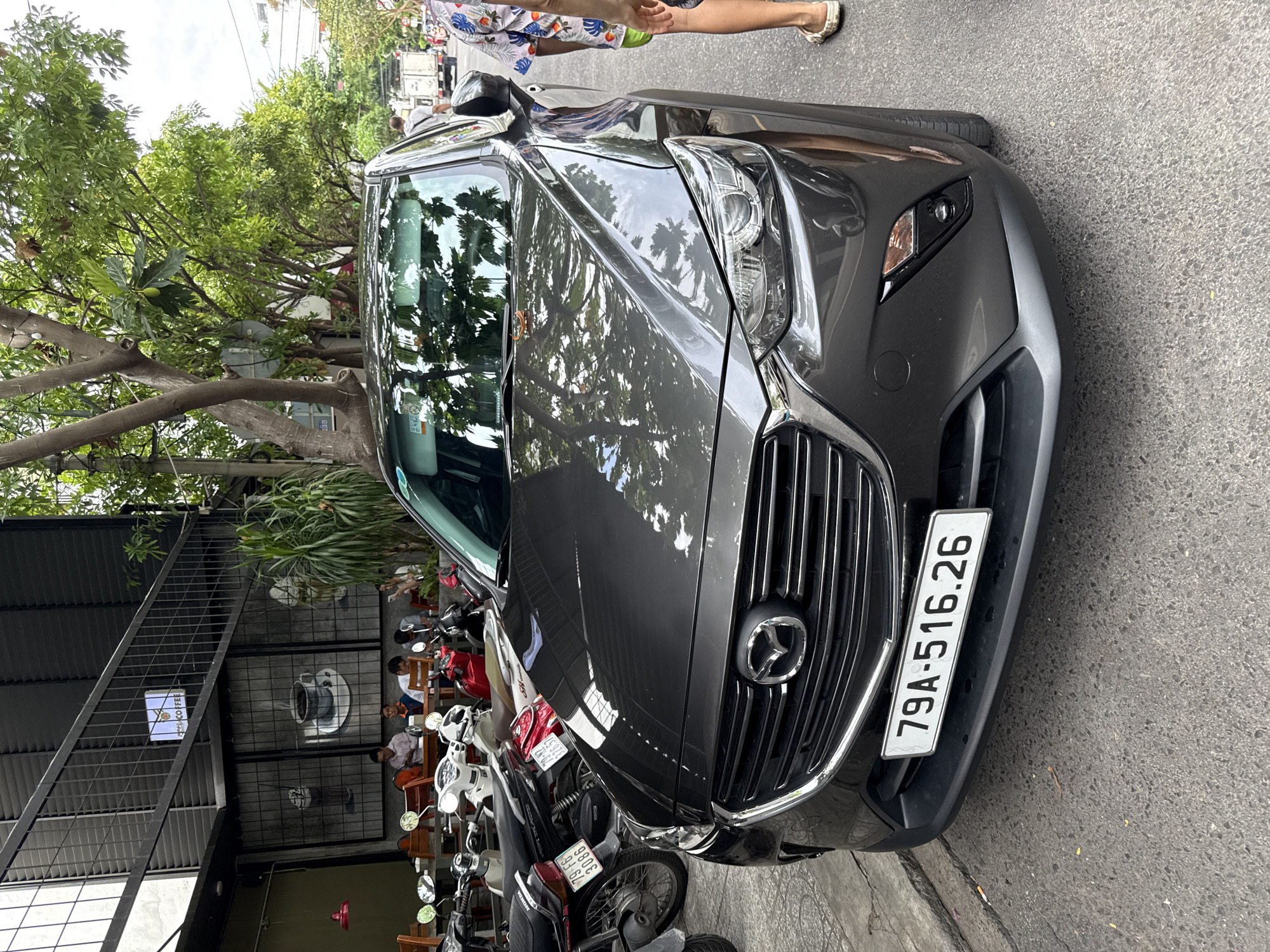 The image shows a dark gray Mazda CX-3 parked on a street in Nha Trang, Vietnam. The car has a license plate that reads "79A-516.26". In the background, there is a coffee shop with outdoor seating and several motorcycles parked. The weather appears to be overcast. A person wearing a brightly colored shirt and green shorts is standing to the right of the car, partially obscuring it from view. The street is paved with asphalt and there is a sidewalk to the left of the car. The scene suggests a typical street view in a Vietnamese city, with cars, motorcycles, and businesses coexisting.