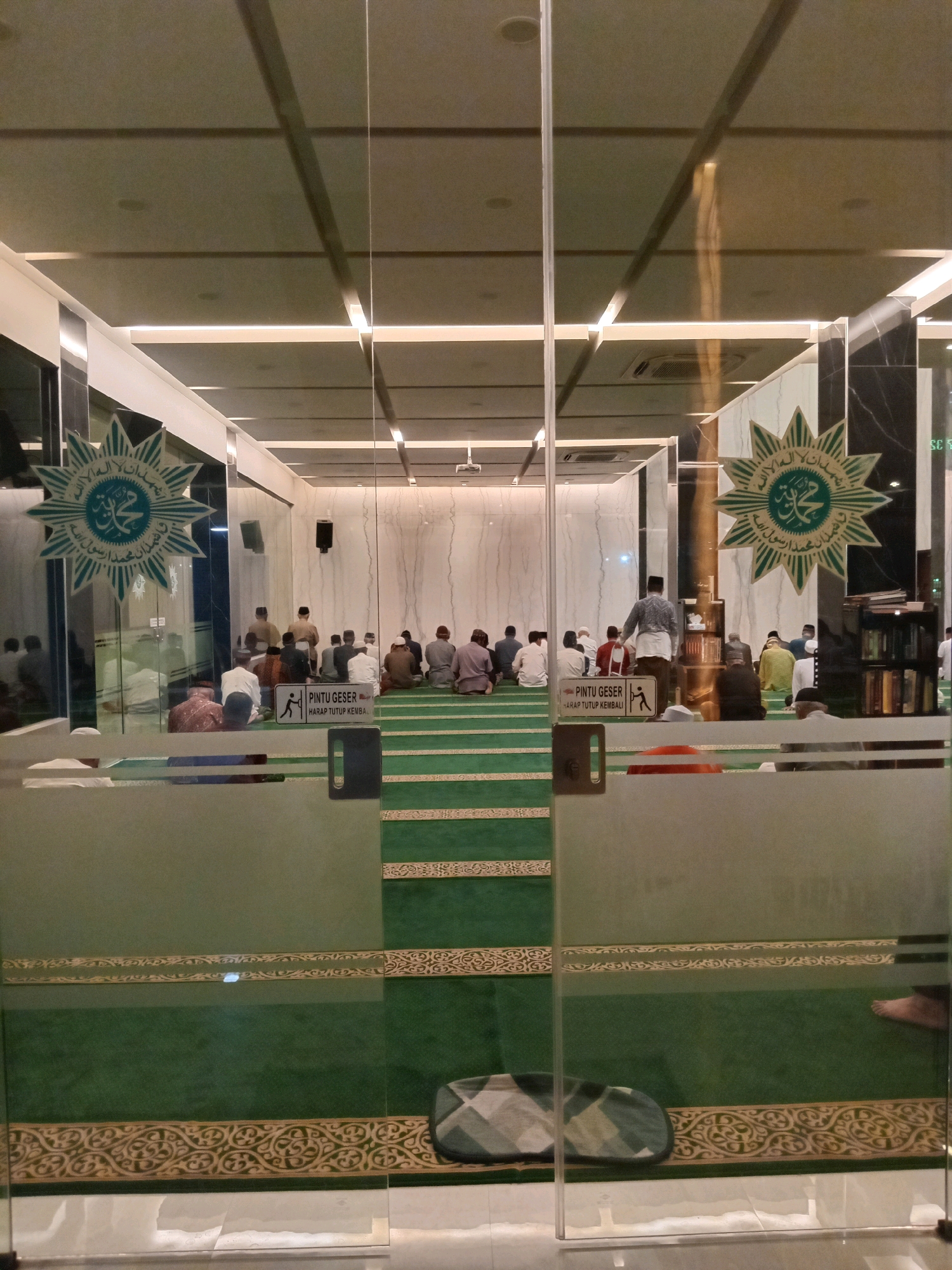 The image shows the interior of a mosque, as seen through a glass door. The mosque has green carpeting with decorative borders, and white walls. Several men are seated on the carpet, facing a wall, appearing to be praying. One man stands near the back of the room, seemingly overseeing the prayer. There are also Islamic symbols on the glass door and on the walls. The lighting appears to be artificial and is integrated into the ceiling. The scene is likely set in Randuagung, Indonesia, given the presence of the text "PINTU GESER HARAP TUTUP KEMBALI" which translates to "Sliding Door Please Close Again" in Indonesian.