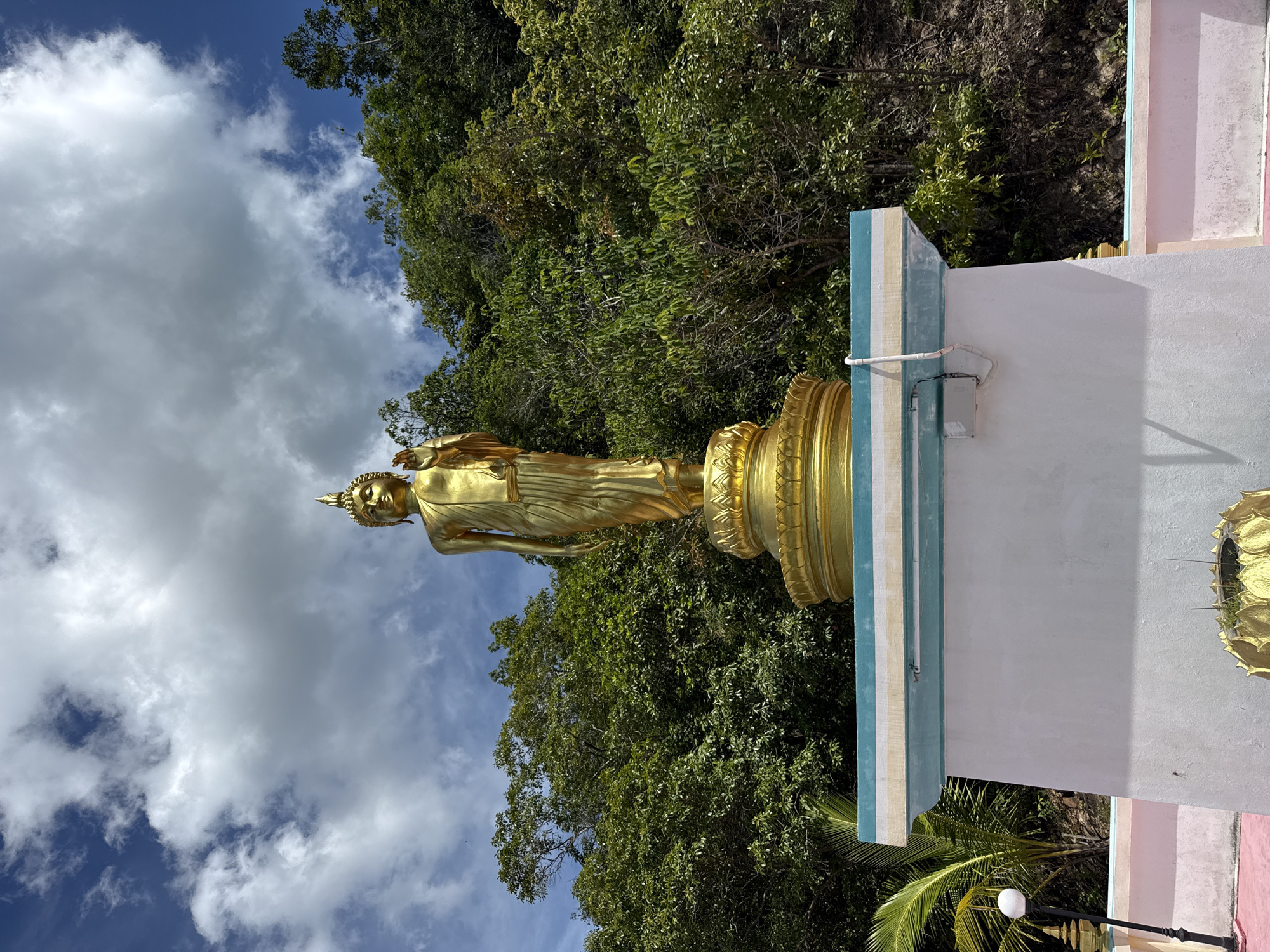 A golden Buddha statue stands on a pedestal in front of a lush green forest under a bright blue sky with fluffy white clouds. The statue is adorned in traditional robes, with one hand raised in a gesture of blessing. The pedestal is decorated with ornate golden lotus petals. The statue is situated on a white platform with blue and yellow trim, which appears to be part of a larger structure, possibly a temple or monument. To the left of the platform, a black lamppost with a white globe is visible, along with a palm tree. The ground is a pale pink color. The scene suggests a serene and spiritual setting in Ko Pha Ngan, Thailand.