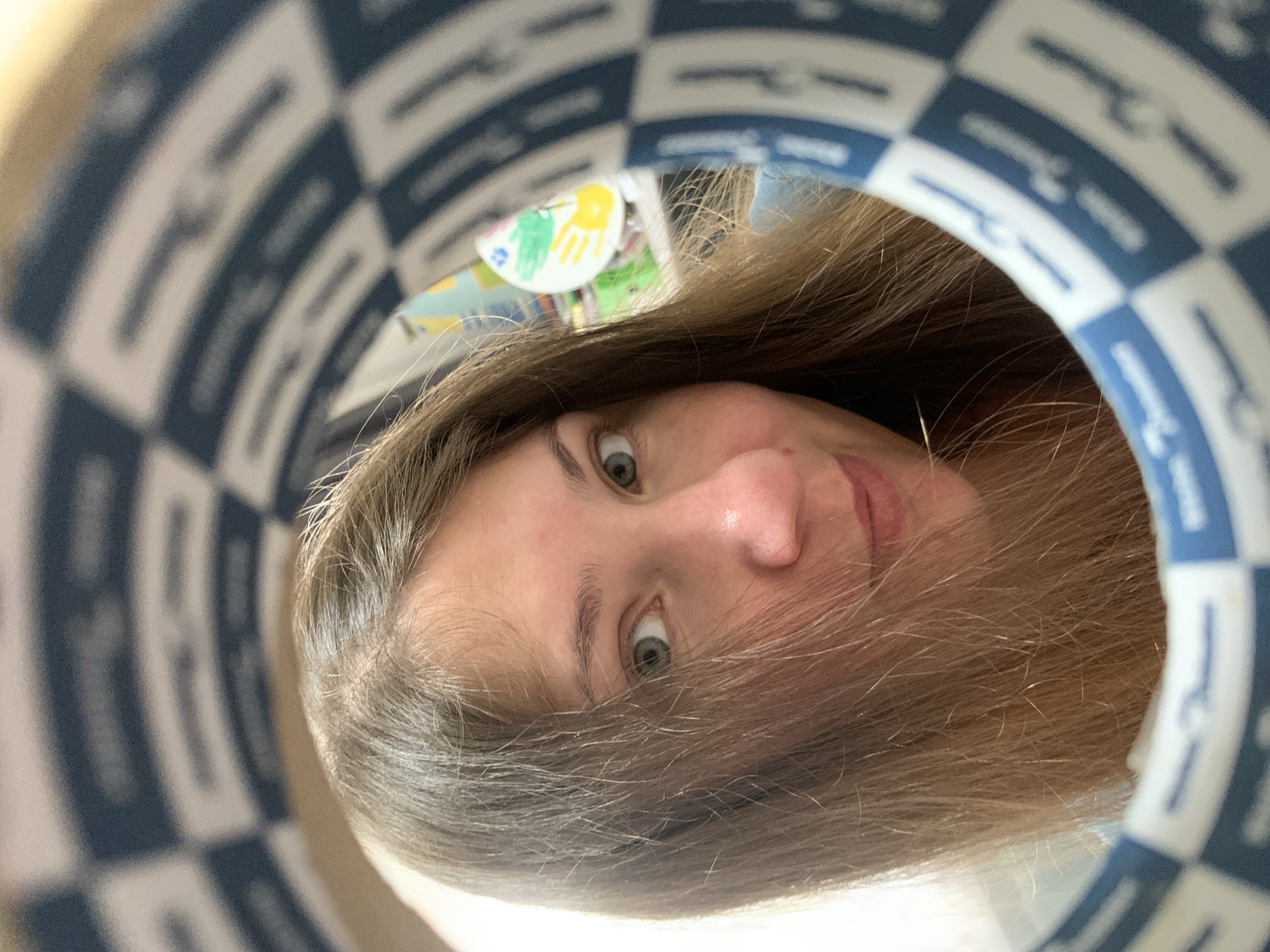 A woman with long brown hair is looking through a circular object that has a blue and white checkered pattern. She is positioned in the center of the frame, with her face taking up a significant portion of the image. Her eyes are blue-green, and she has a light complexion. Her hair is slightly covering her face. In the background, out of focus, there are some colorful objects which appear to be children's toys or decorations. The circular object she is looking through is partially obscuring her face, creating a tunnel-like effect. The image is a close-up, portrait-style photograph. The scene appears to be indoors, in what could be a domestic or playful environment. There is no discernible information in the image to indicate the specific location in Chaikovsky, Russia.