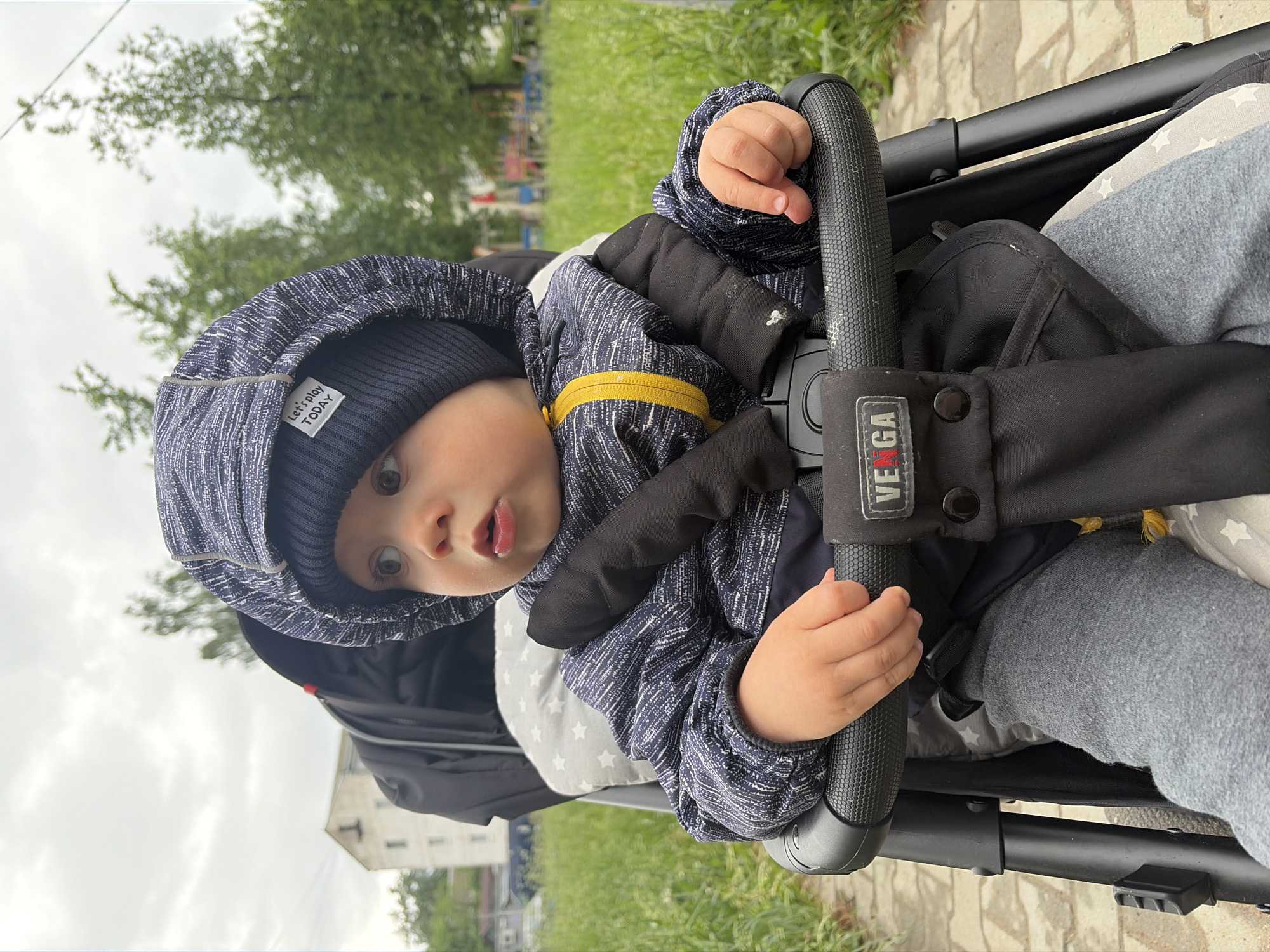 A baby sits in a stroller outside. The child wears a blue hat with a "Let's play TODAY" tag and a matching hooded jacket over black padding with a yellow zipper. The stroller has a "VEGA" logo. The location is in Chaikovsky, Russia. The baby is touching the stroller's handle. Grass and a building are visible in the background.