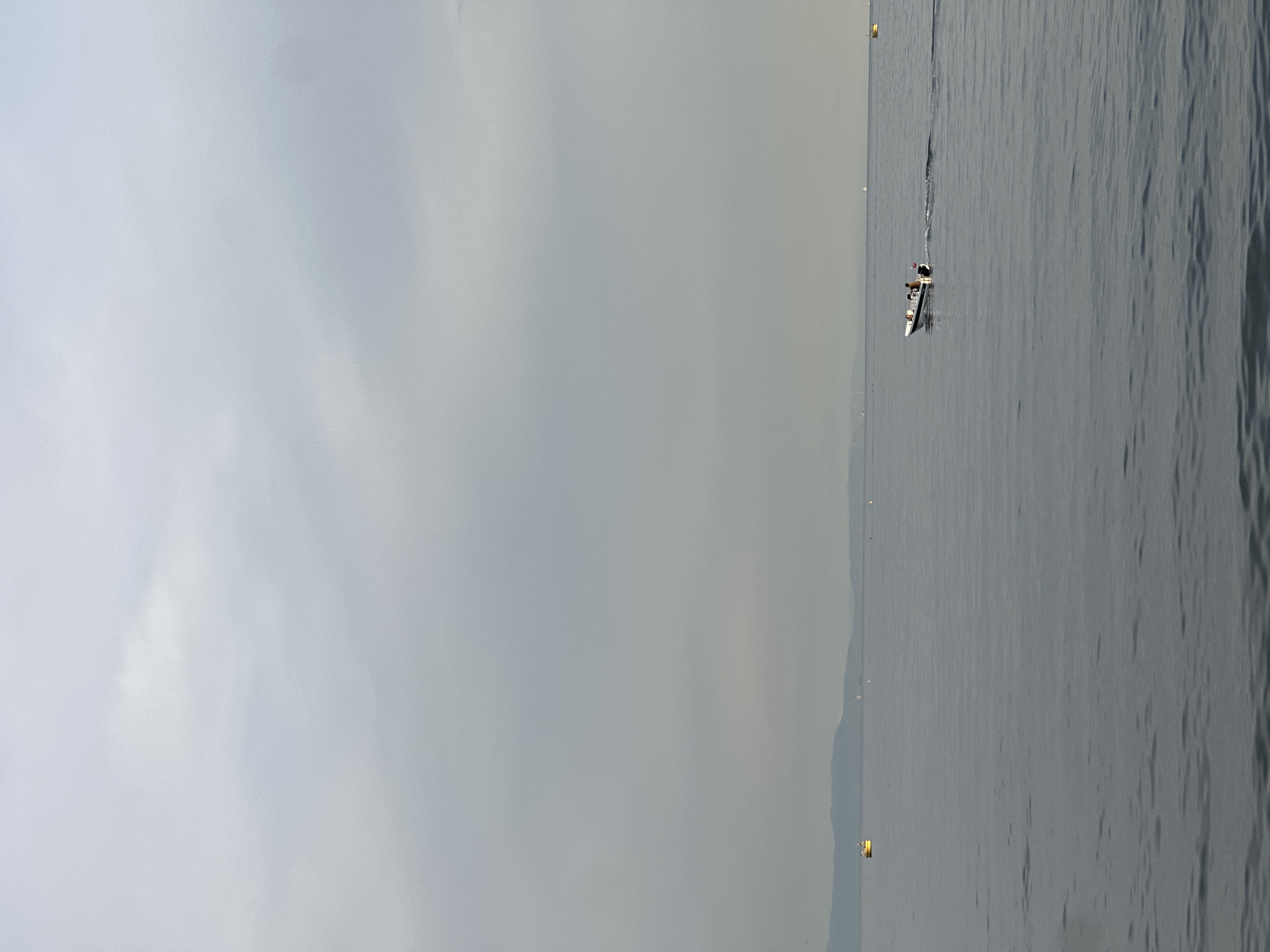 A wide shot captures a hazy, overcast day over the Sea of Marmara, likely near Bakırköy, Turkey. The water is calm and a muted gray, with subtle ripples on the surface. In the middle distance, a small white motorboat with at least two figures aboard moves from left to right, leaving a small wake. Further to the left, a bright yellow buoy floats on the water. On the horizon, a faint, low-lying landmass with rolling hills or mountains is visible, possibly the coastline near Bakırköy. Several other distant, small white objects, possibly buoys or distant boats, are scattered across the water. The sky is a uniform pale gray, with diffused light indicating a lack of direct sunlight. The overall atmosphere is serene and slightly muted due to the overcast conditions.