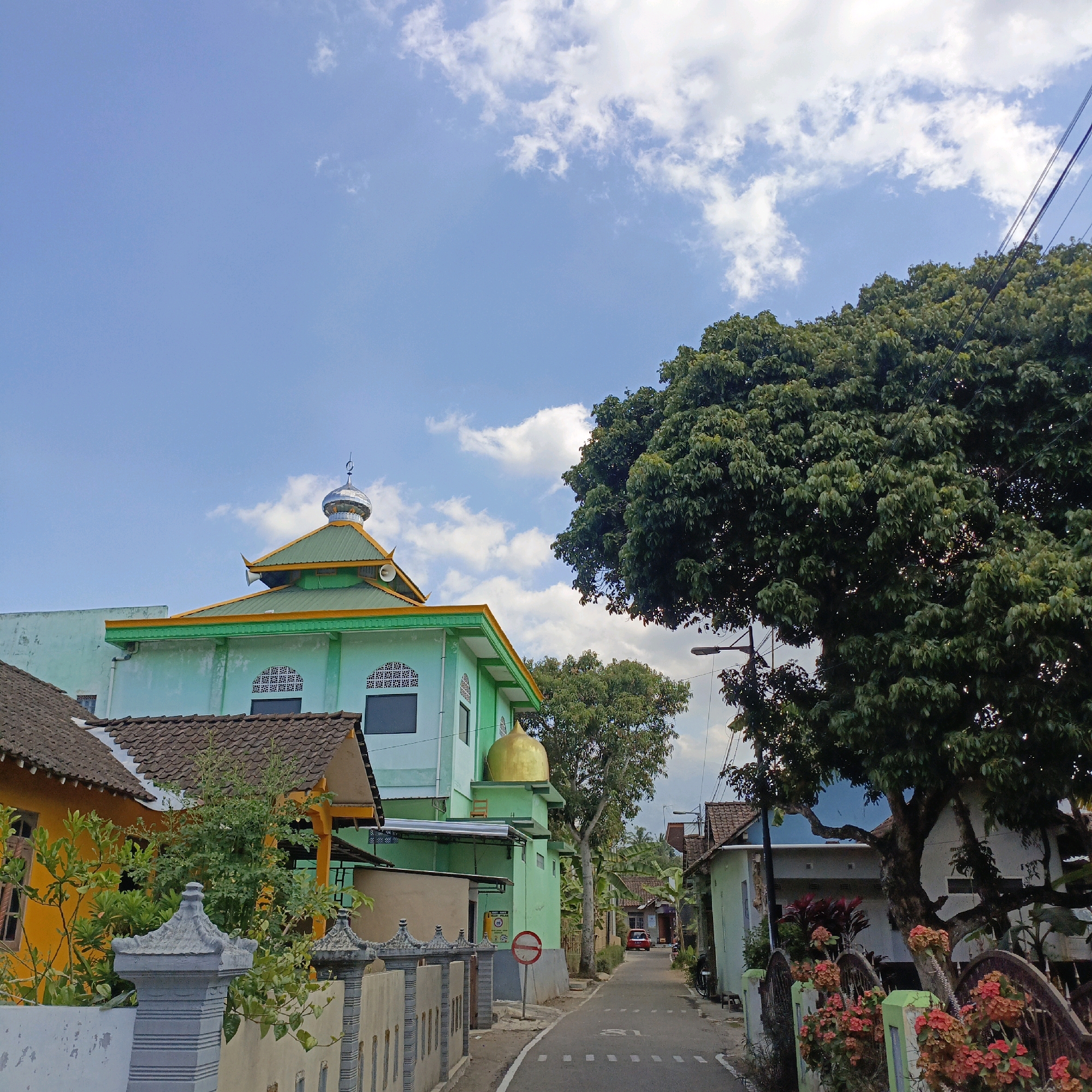 This is a street scene in Salatiga, Indonesia. A light blue sky with scattered white clouds dominates the upper portion of the image. In the mid-ground, a predominantly green mosque with a golden dome is visible on the left. The mosque has a prominent silver dome topped with a crescent moon and star symbol. Adjacent to the mosque, a yellow house with a brown tiled roof is partially visible. A paved road runs through the center of the scene, lined with various houses and lush green trees. On the right side of the road, a large, mature tree with dense foliage overshadows some of the houses. In the distance, a red car is parked on the side of the road. The foreground features a grey decorative fence and gate. The overall impression is a quiet residential street in a tropical climate.