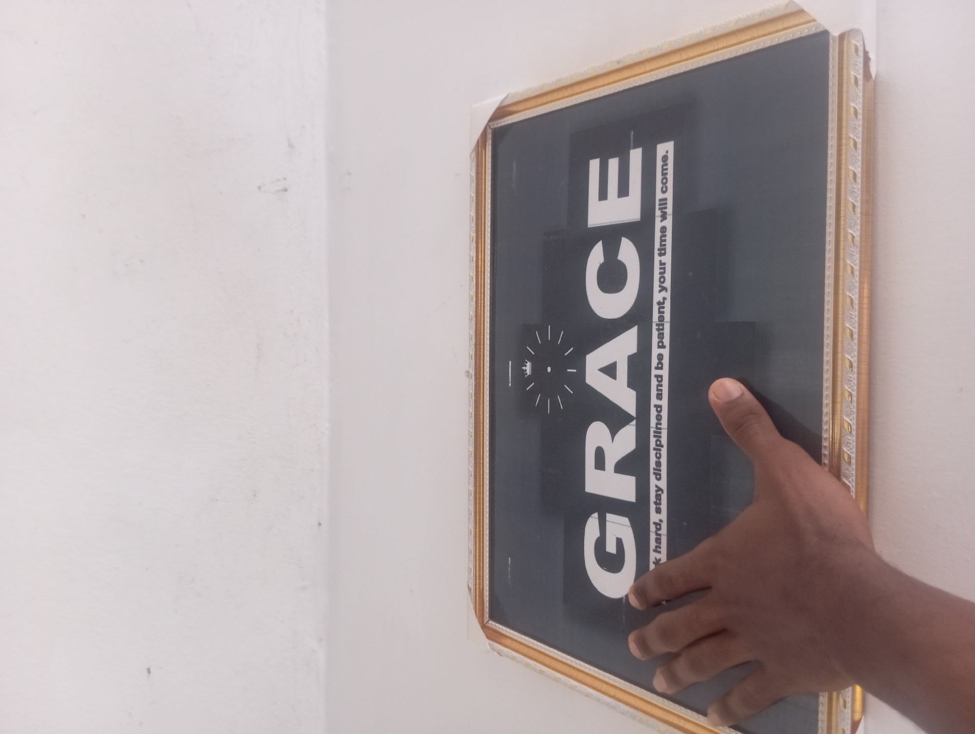 A dark-skinned hand is partially visible on the right side of the frame, touching or holding a framed picture against a plain white wall. The picture features a black background with bold white text. The prominent word "GRACE" is displayed in large capital letters. Below it, smaller text reads: "Work hard, stay disciplined and be patient, your time will come." Above the "R" in "GRACE," there is a small graphic resembling a sunburst or a stylized clock face. The picture is encased in a golden-colored frame with visible decorative patterns along its edges. The setting appears to be indoors, with the corner of two white walls visible towards the left side of the image.
