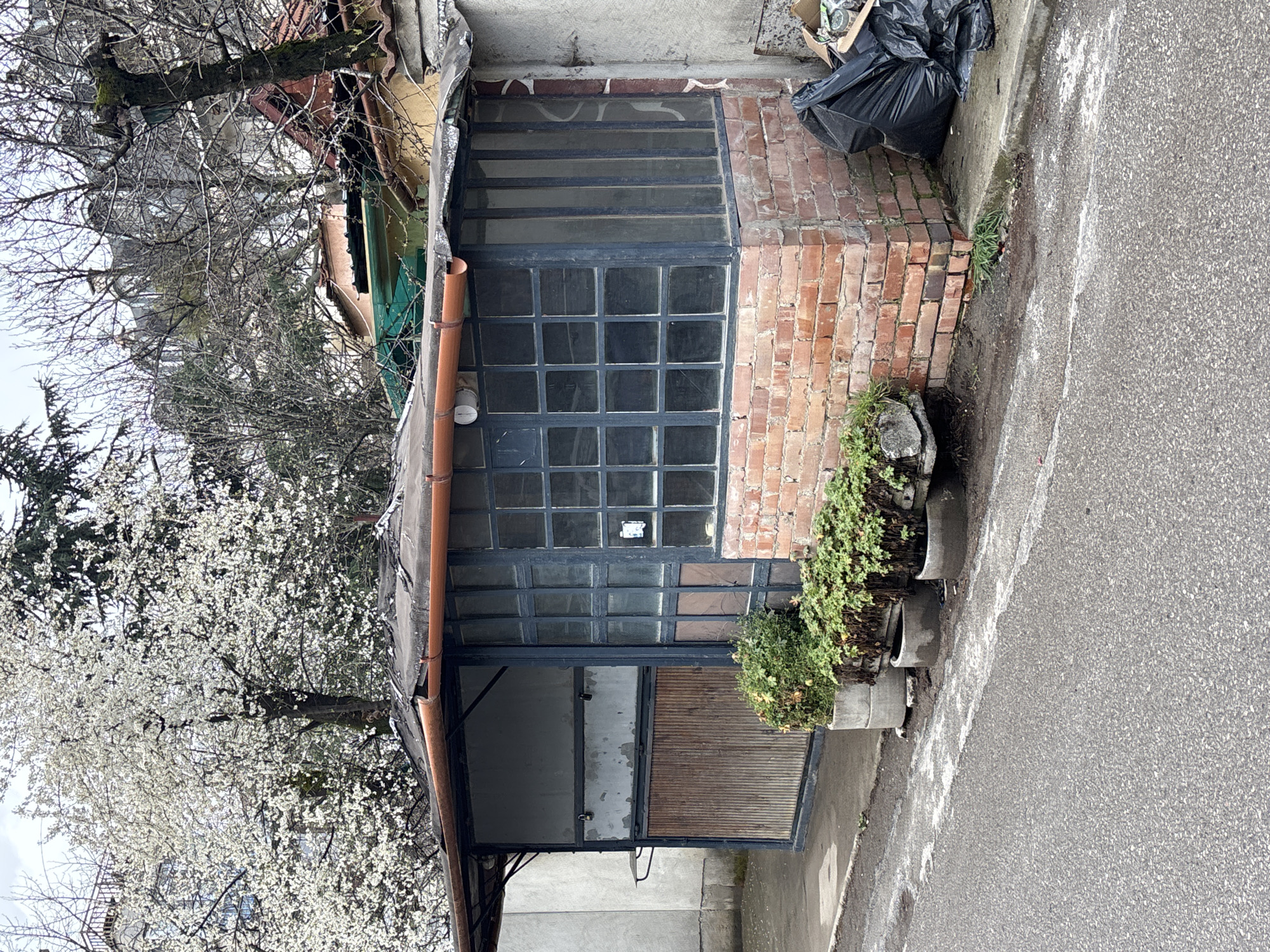 The image depicts a small, dilapidated structure in Sofia, Bulgaria. The structure appears to be an old garage or storage shed, with a brick wall on the right and a large window with multiple panes. The window is dirty and has some broken panes. A black trash bag sits on the roof. An orange rain gutter is attached to the roof. To the left, a tree with white blossoms is in full bloom. A narrow street borders the structure on the right. The overall impression is one of neglect and disrepair.