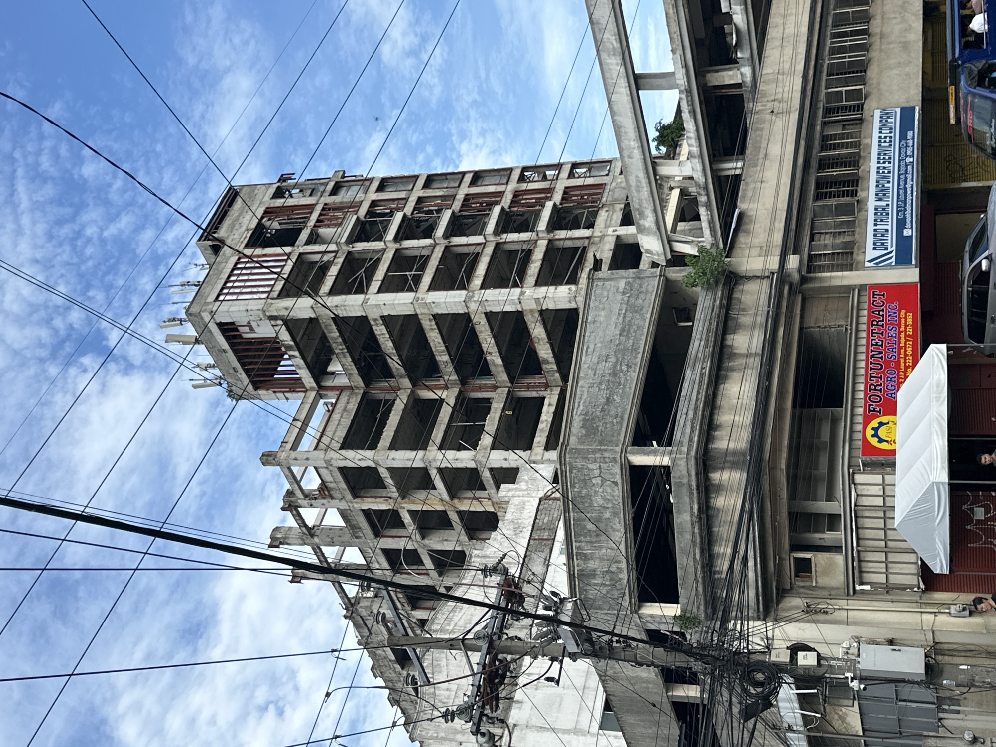 The image shows a street scene in Davao City, Philippines. An unfinished concrete building stands prominently, its skeleton-like structure exposed. Adjacent to it is a more complete building with commercial signage, including "FORTUNETRACT AGRO-SALES INC." and "DAVAD TRIBAL MANPOWER SERVICES COMPANY." Power lines densely crisscross the view. A few people are visible in the background near the commercial building. The sky is partly cloudy.