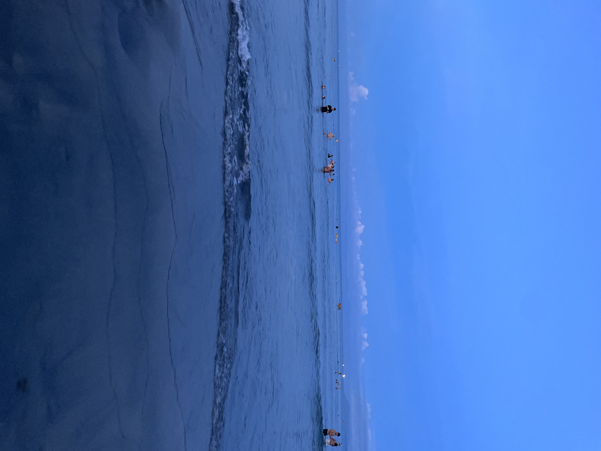 The image depicts a twilight scene at a beach in Da Nang, Vietnam, likely My Khe Beach given the popularity and typical scenery. The sky is a deep, clear blue with a few wispy white clouds near the horizon. The sea is calm with gentle waves lapping at the shore. Several people are visible in the water, some wading, others swimming, and a few appear to be standing or playing. In the distance, there are faint lights, possibly from other beachgoers or structures. The foreground shows the wet sand reflecting the dark blue of the sea and sky. The overall atmosphere is serene, with the natural beauty of the beach illuminated by the fading daylight.