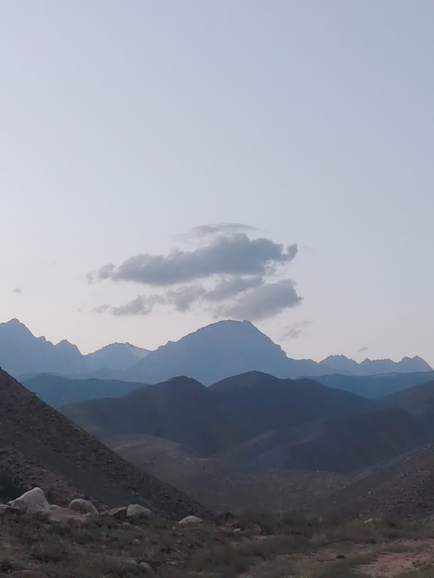 This is a landscape photograph taken in the city of Cholpon-Ata, Kyrgyzstan, featuring a mountain range under a clear, pale blue sky. The sky occupies the upper portion of the image and transitions to a lighter hue near the horizon. A single, wispy cloud formation is visible in the middle ground. The mountains are layered, with each range appearing progressively darker and more distant. The closest mountains in the foreground are rugged and appear to be covered in sparse vegetation and rocks, with some larger boulders scattered across the terrain. The middle ground and background consist of soft, undulating mountain slopes that recede into the distance, creating a sense of depth. The lighting suggests it might be early morning or late evening, with soft, diffused light casting subtle shadows. There are no people or animals visible in the image. The scene is serene and natural, emphasizing the grandeur of the mountainous landscape.