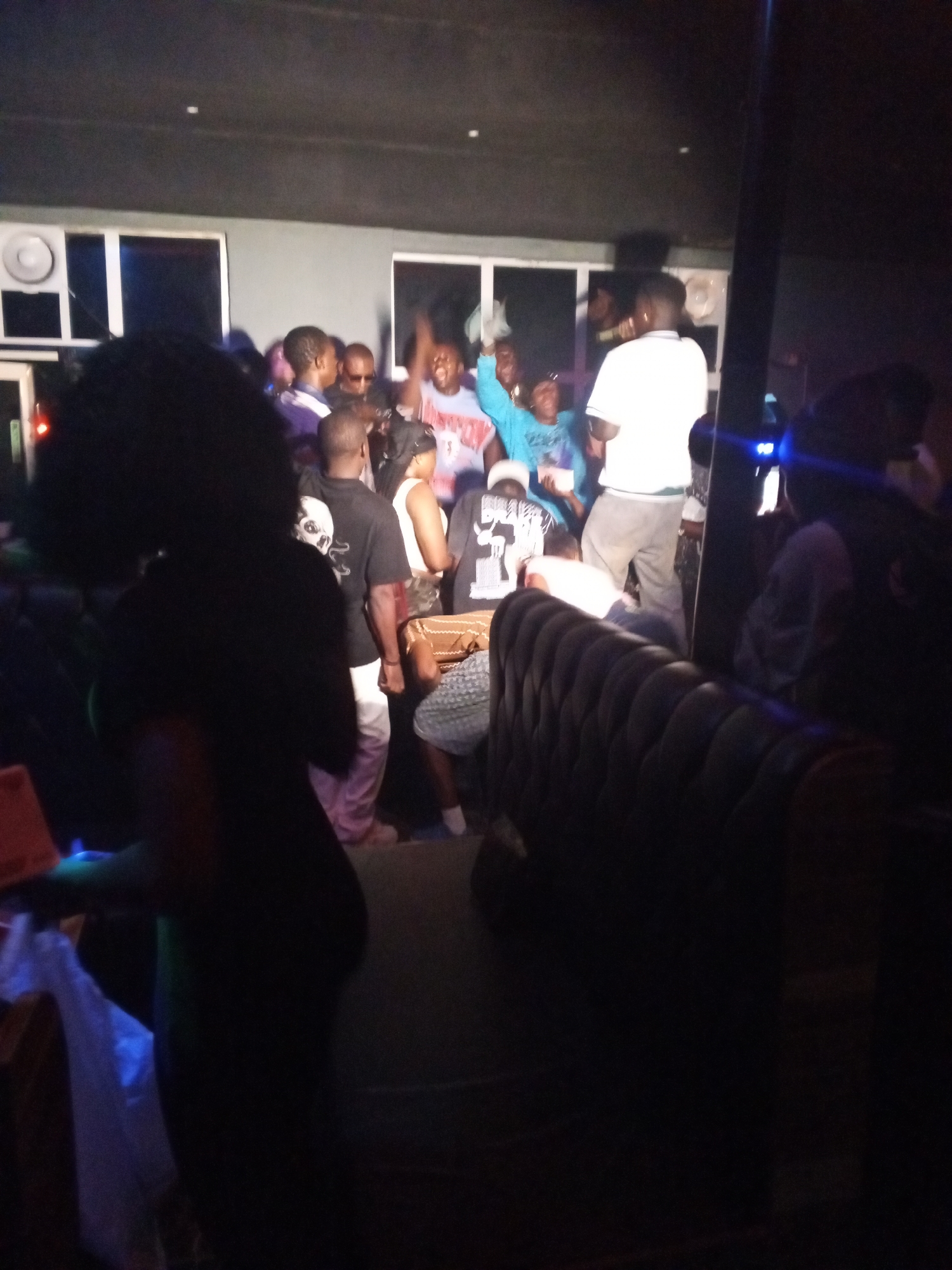 A dark, grainy photograph shows a group of people gathered in a dimly lit indoor space, likely a nightclub or lounge. The location may be in Lokoja, Nigeria. People are standing, some with arms raised, suggesting an active or celebratory atmosphere. The room has a dark-colored wall with visible speakers and window-like structures. A dark leather-like tufted booth is visible in the foreground, indicating seating within the space. The overall image quality is poor, making details difficult to discern.