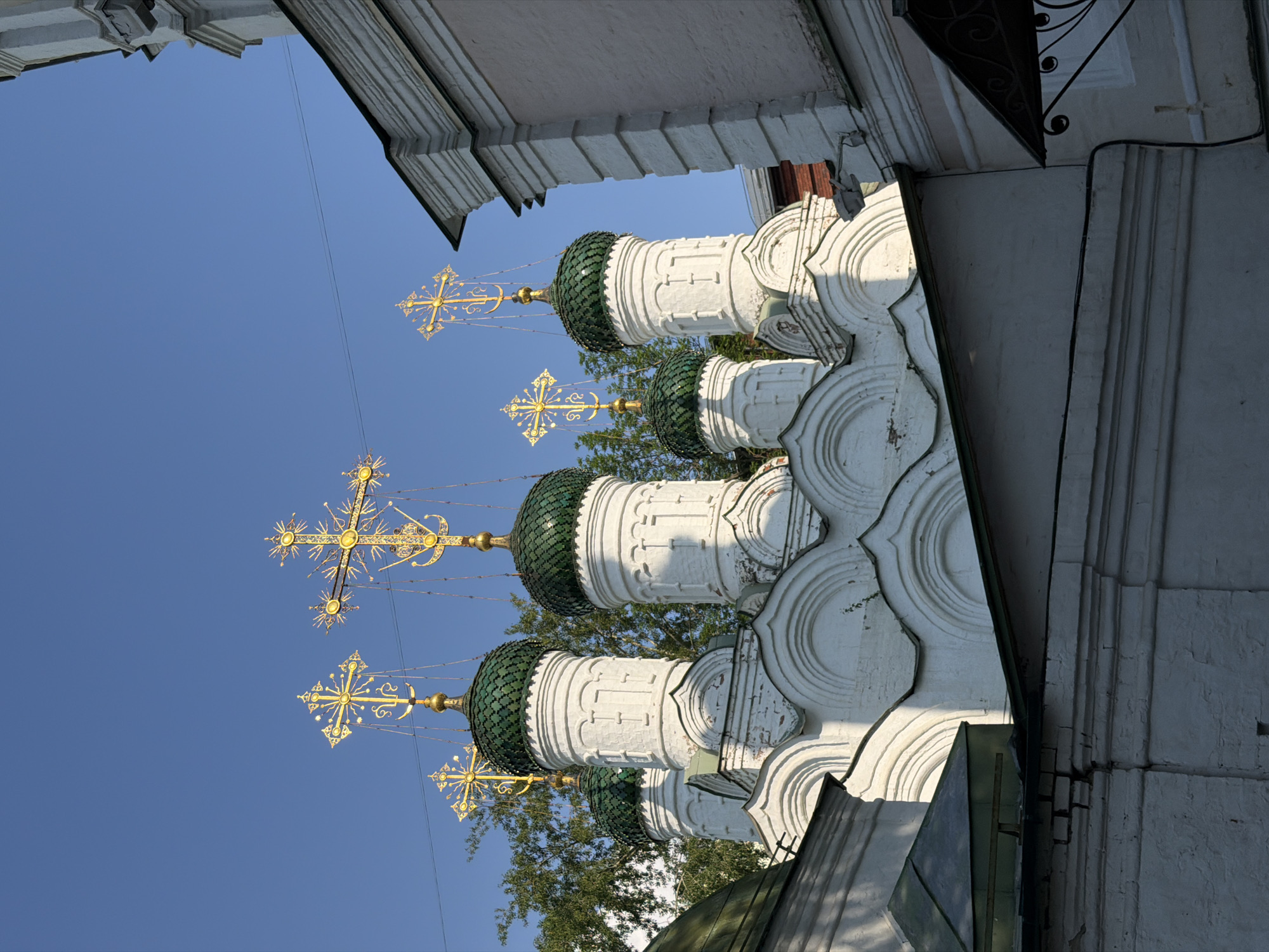 The image shows a low-angle view of the domes and crosses of a Russian Orthodox church, likely located in Moscow, Russia. The church features white walls with decorative, semi-circular patterns and green, onion-shaped domes. Golden crosses with elaborate starburst designs are mounted atop each dome. The sky is a clear blue. Architectural details of the church and a neighboring building are visible along the edges of the frame. Trees can be seen behind the domes.