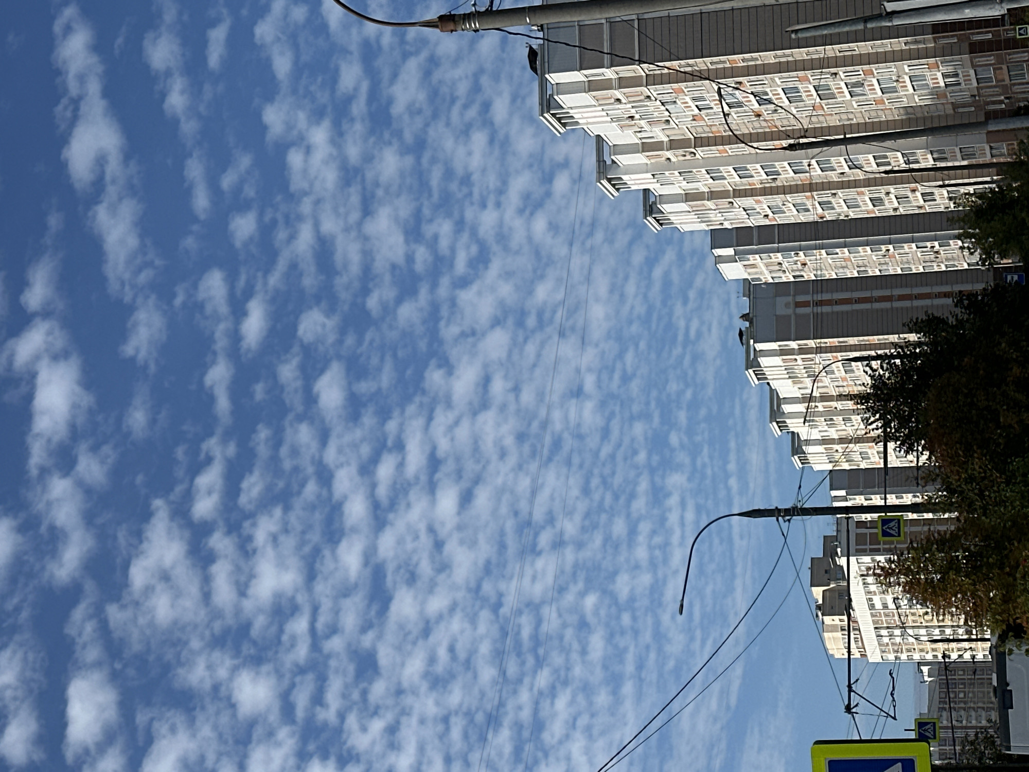 A low-angle, full shot captures a row of residential buildings against a bright blue sky filled with small, scattered white clouds. The buildings, which appear to be multi-story apartment complexes, are white and light gray with orange accents and numerous windows. They are aligned diagonally from the lower right to the upper left of the frame, creating a sense of depth. In the foreground, electrical wires and streetlights crisscross the scene, and a pedestrian crossing sign is visible on the left. Trees with autumn foliage are interspersed between the buildings and in the foreground. The environment appears to be an urban setting in Podolsk, Russia, likely during the daytime due to the bright sky and the shadows cast. There are no people visible in the image.