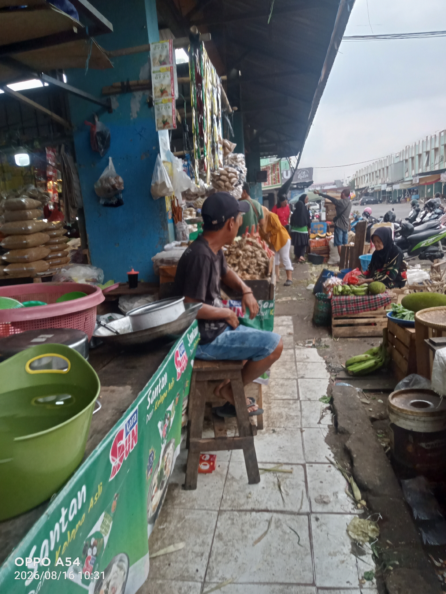 This is a street-level, medium shot photograph taken outdoors in the city of Jambi, Indonesia, likely a market or roadside stall. The image is taken from a slightly low angle and appears to be a candid shot.
In the foreground, a green plastic bucket filled with water is visible on the left side. To the right of the bucket, a long, green banner with Indonesian text and illustrations is displayed. Part of the banner reads "Santan Kelapa Asli". Below the banner, a man in a black t-shirt and blue shorts is seated on a wooden stool. He is looking towards the right of the frame and has his left arm resting on his leg. His facial features are not clearly visible.
Behind the man, there are stalls with various goods. To the left, stacks of brown bags filled with what appear to be grains or spices are piled up. In the center, there are metal bowls and other utensils. Further back, other people are visible. A woman in a black headscarf and patterned clothing is seated behind a display of green vegetables, including what appears to be bananas and a large jackfruit. Other individuals are standing and walking around, some interacting with vendors.
The environment suggests a busy marketplace. The stalls are constructed with a mix of materials, including wood and metal. The ground is paved with light-colored tiles. In the background, there are buildings and a street with parked motorcycles and some vehicles, indicating an urban setting. The sky is overcast. The overall impression is one of a typical day at a local market in Jambi.