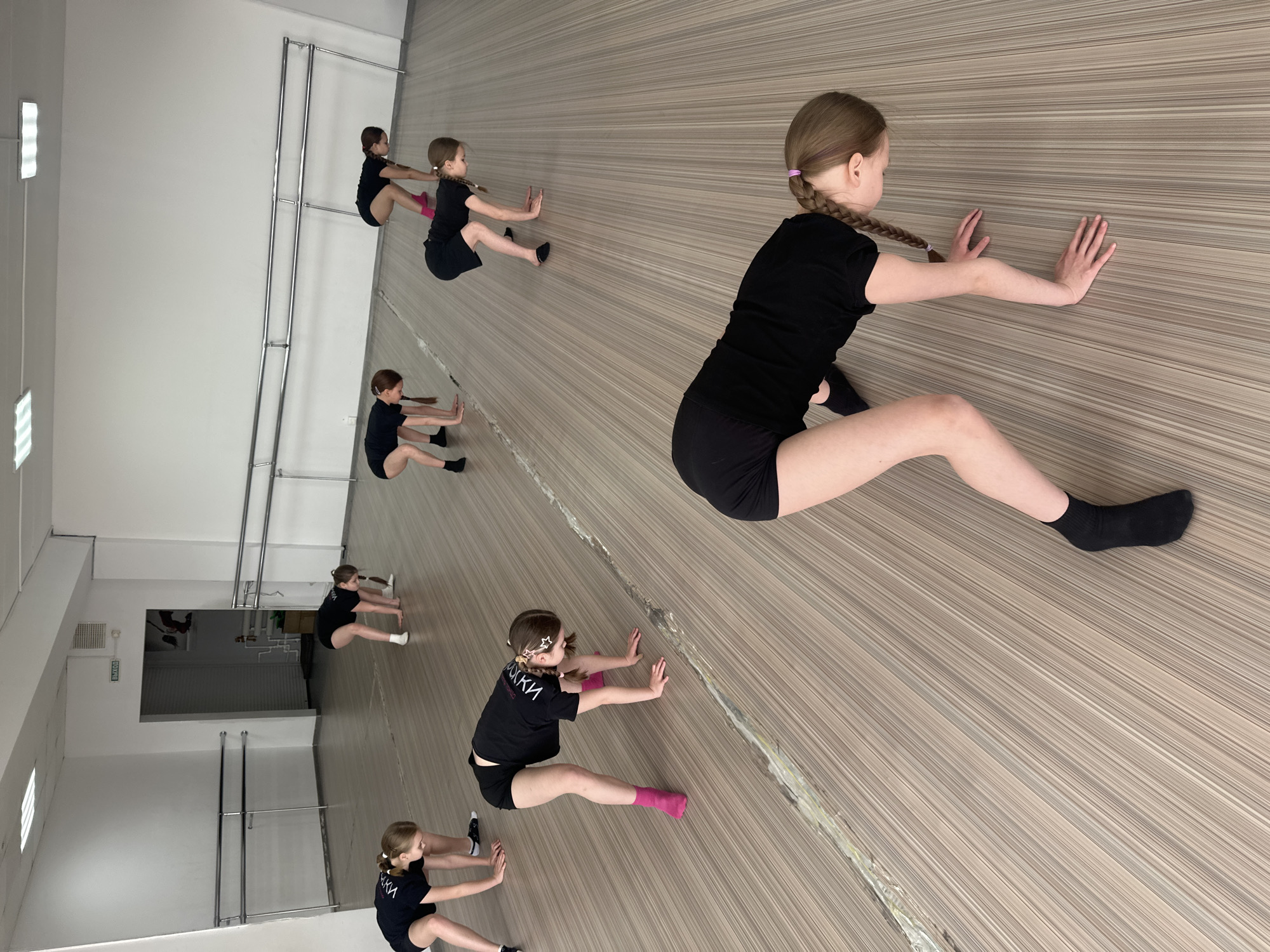 A group of young girls are practicing ballet in a studio in Votkinsk, Russia. They are all wearing black leotards and shorts and have their hair in braids. They are squatting against a wall with their hands placed flat on the surface for balance. The studio has a barre along one wall and a mirror along another. The floor is made of a light-colored wood.